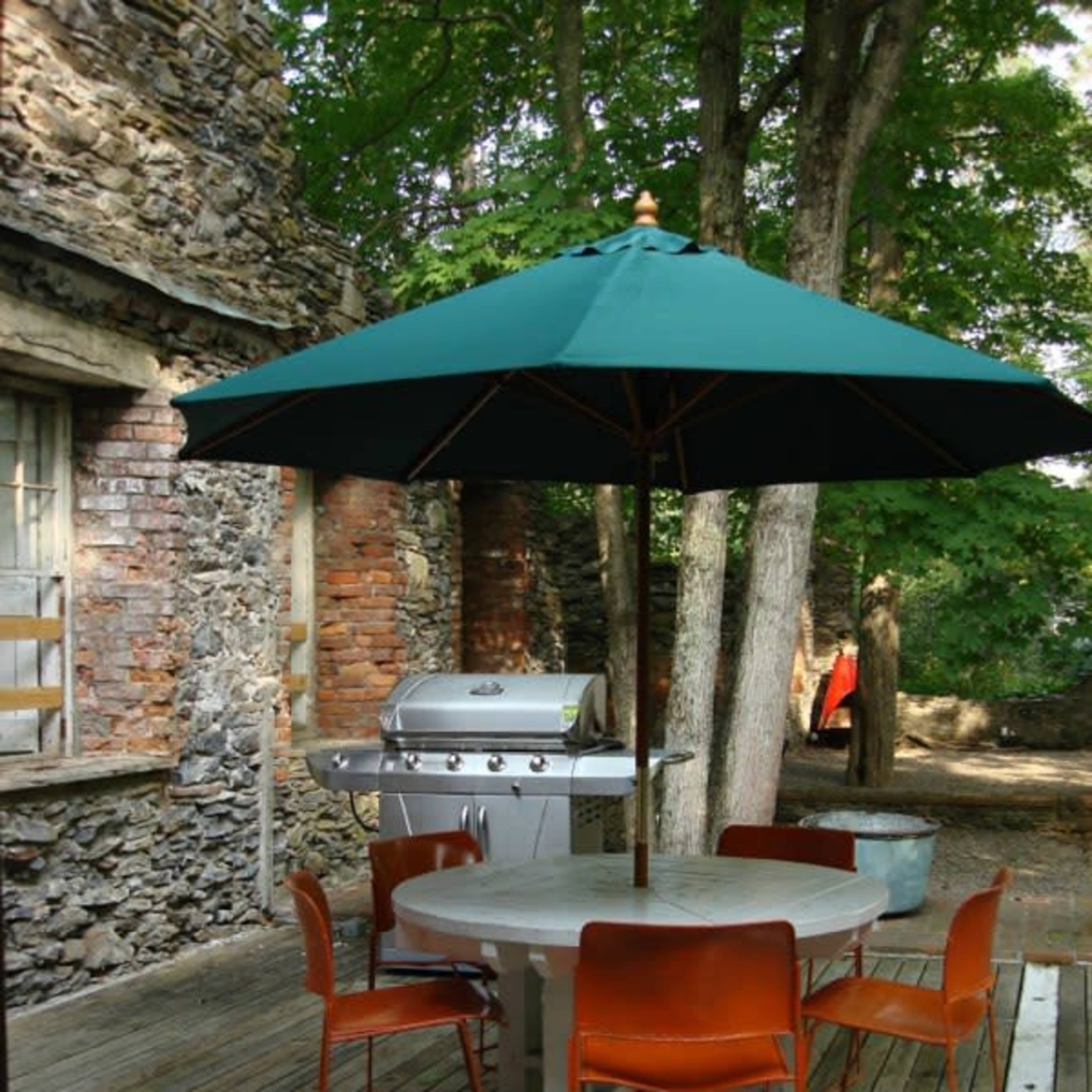 A round patio table with orange chairs and a green umbrella is set on a wooden deck beside a stone wall and a grill.
