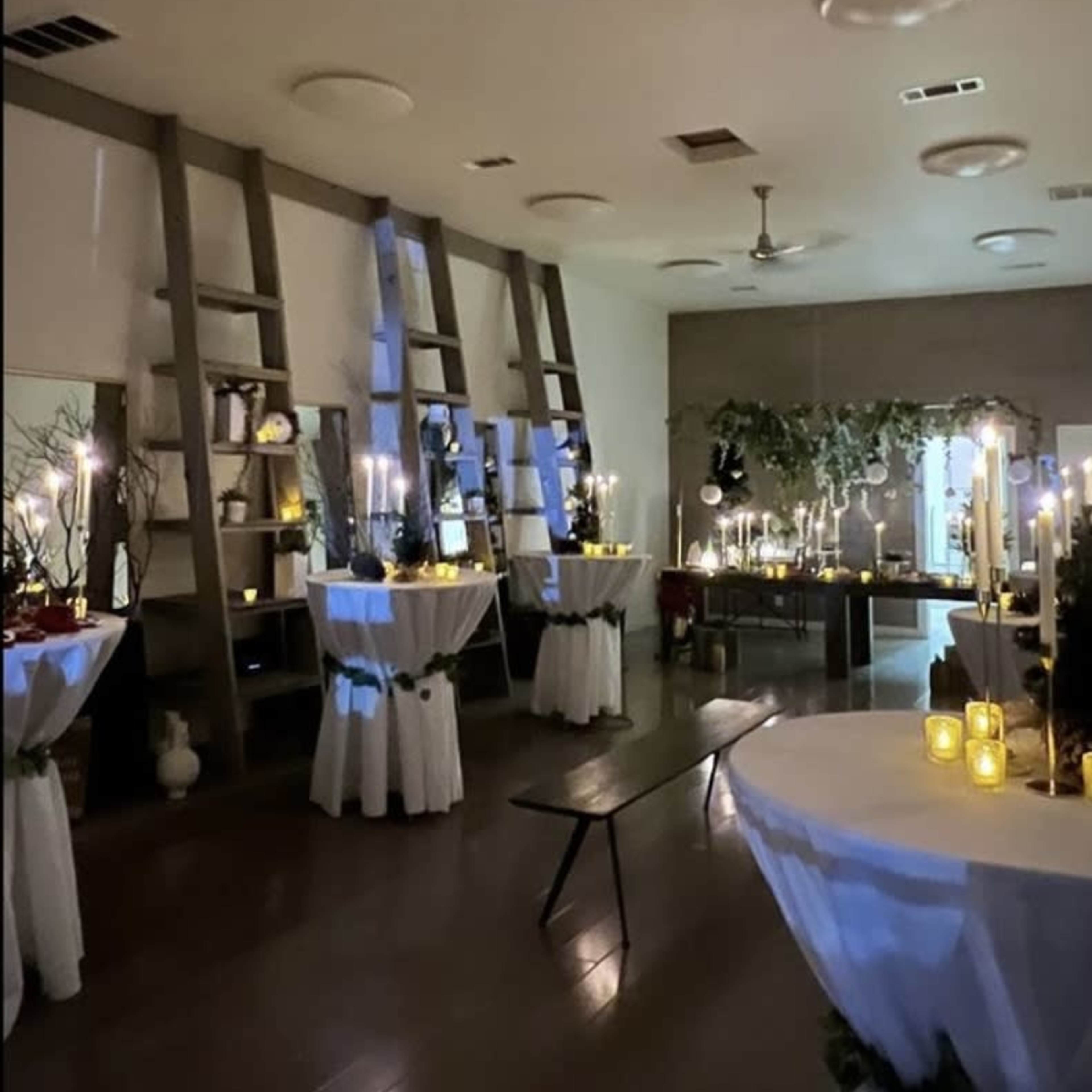 The scene depicts a dimly lit event space with several round tables draped in white linen, surrounded by candles and decorative elements on display shelves.