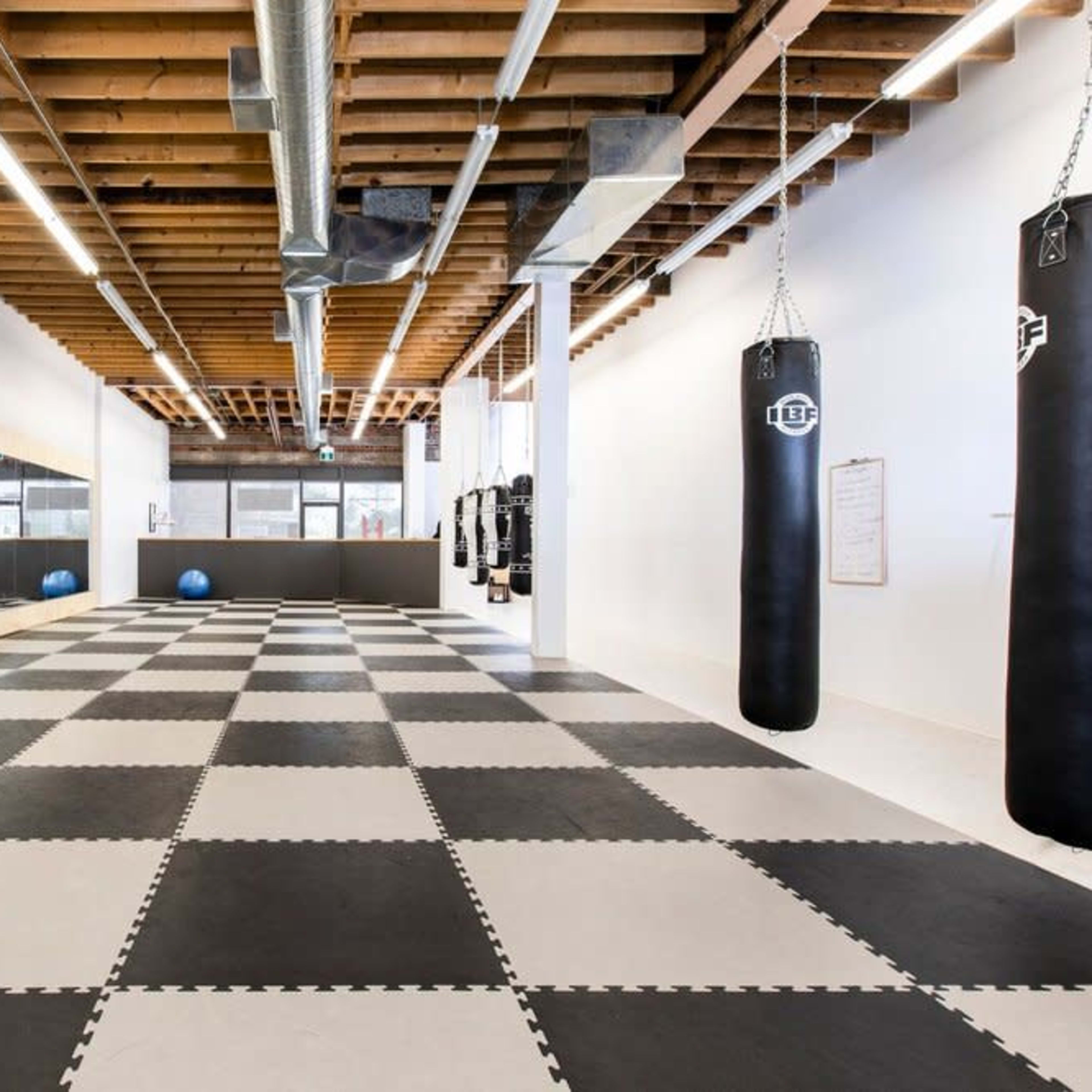 A spacious, empty training area with black and white checkered flooring, reflective mirrors on one wall, and several hanging punching bags.