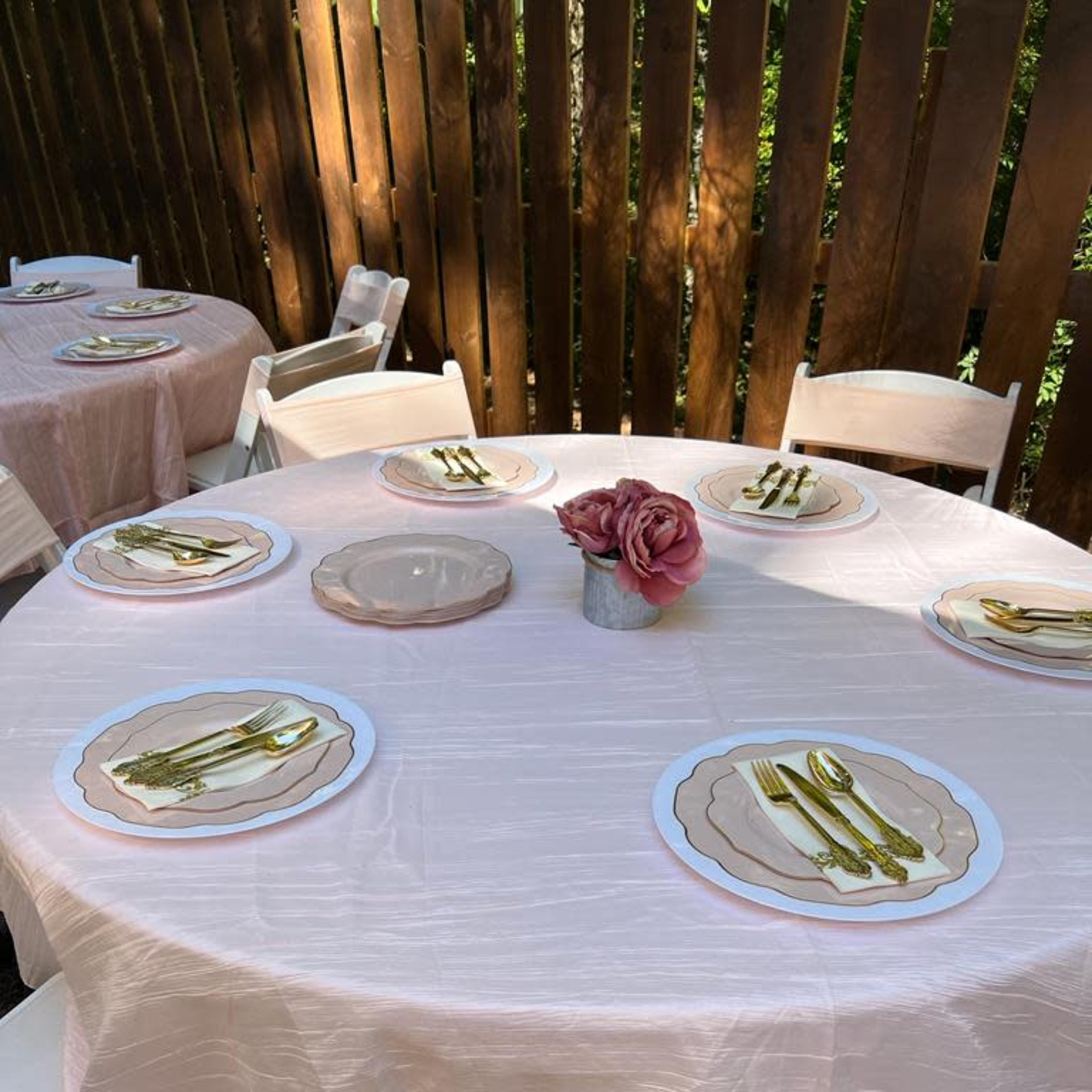 A set of round tables covered with pink tablecloths, each arranged with plates and cutlery, sits against a backdrop of wooden fencing.