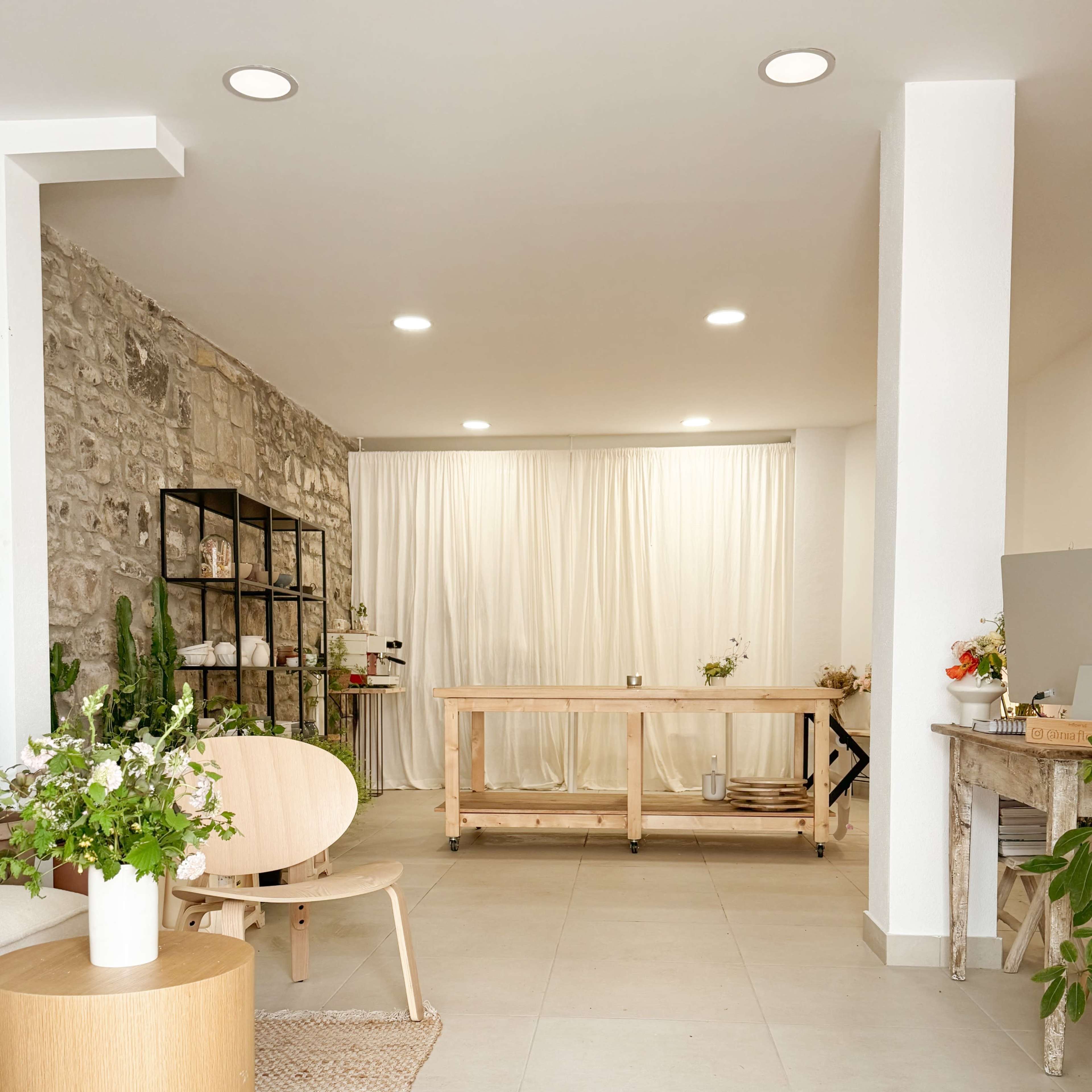 A minimalist interior space with a wooden table, light-colored flooring, and a stone wall, featuring plants and soft curtains in the background.