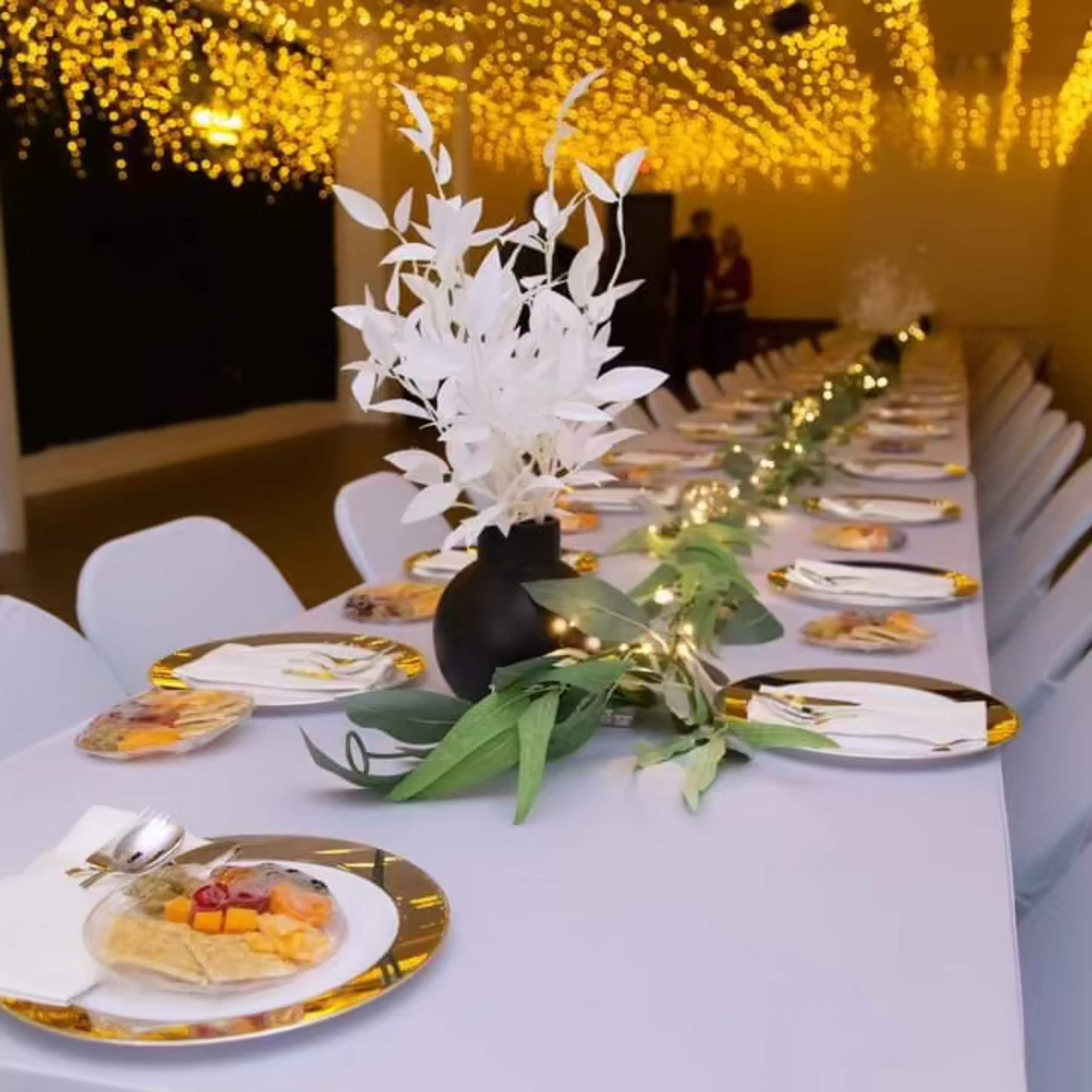 A long dining table is elegantly set with white plates and a decorative vase of white foliage, surrounded by soft lighting from overhead strings of lights.