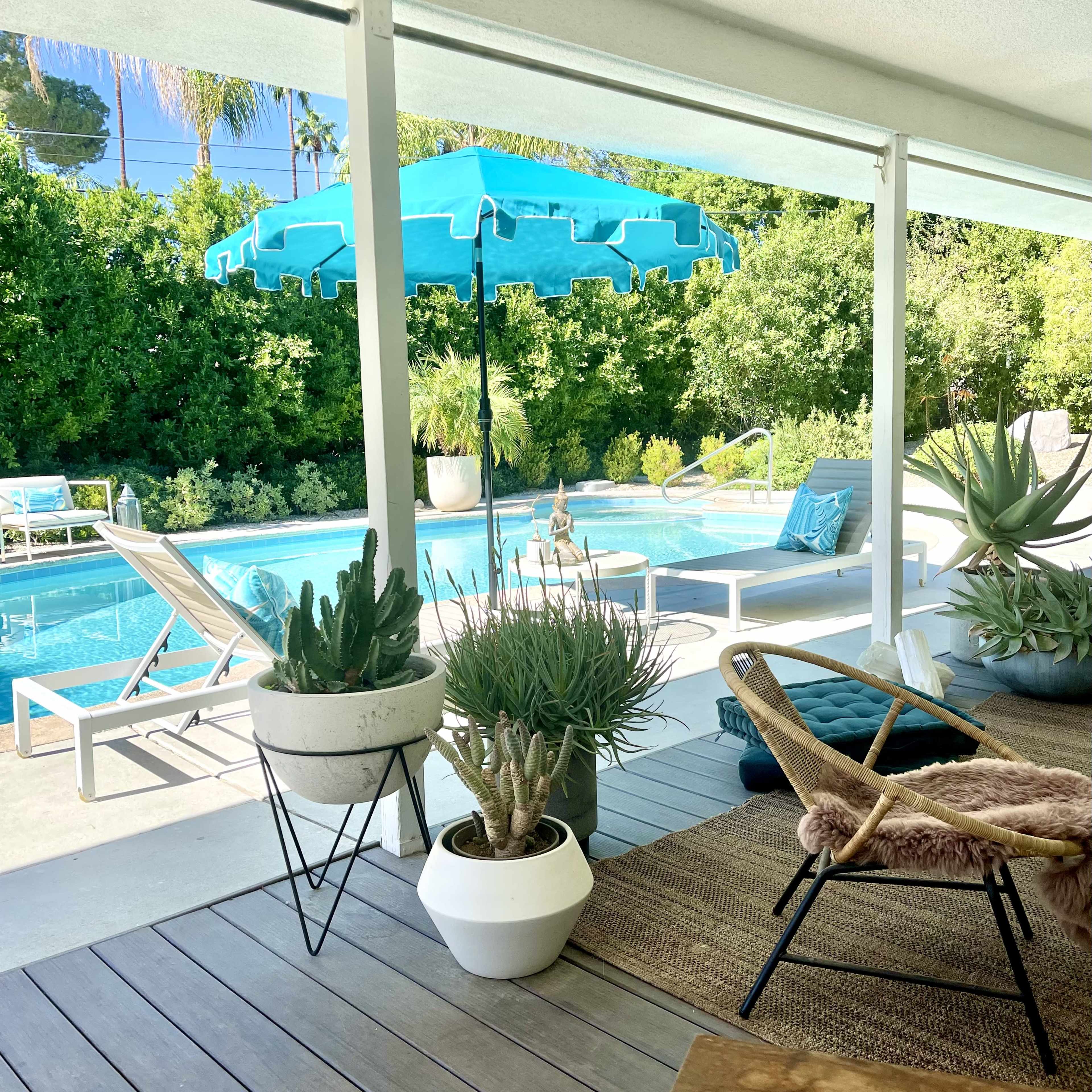 A covered patio overlooks a swimming pool surrounded by greenery, with potted plants and lounge chairs arranged nearby.