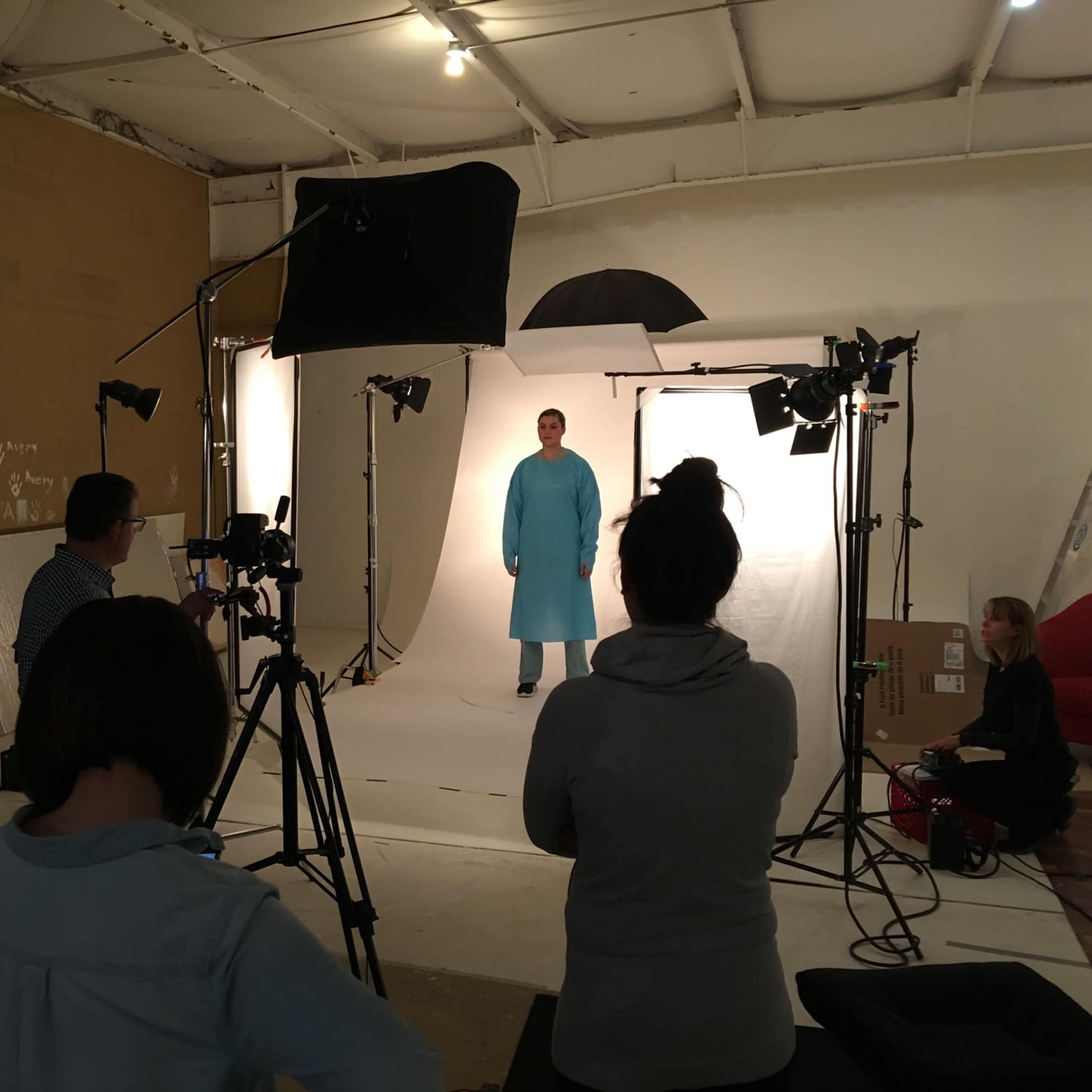 A photoshoot is taking place in a studio, featuring a model in a blue outfit surrounded by multiple cameras and lighting equipment.