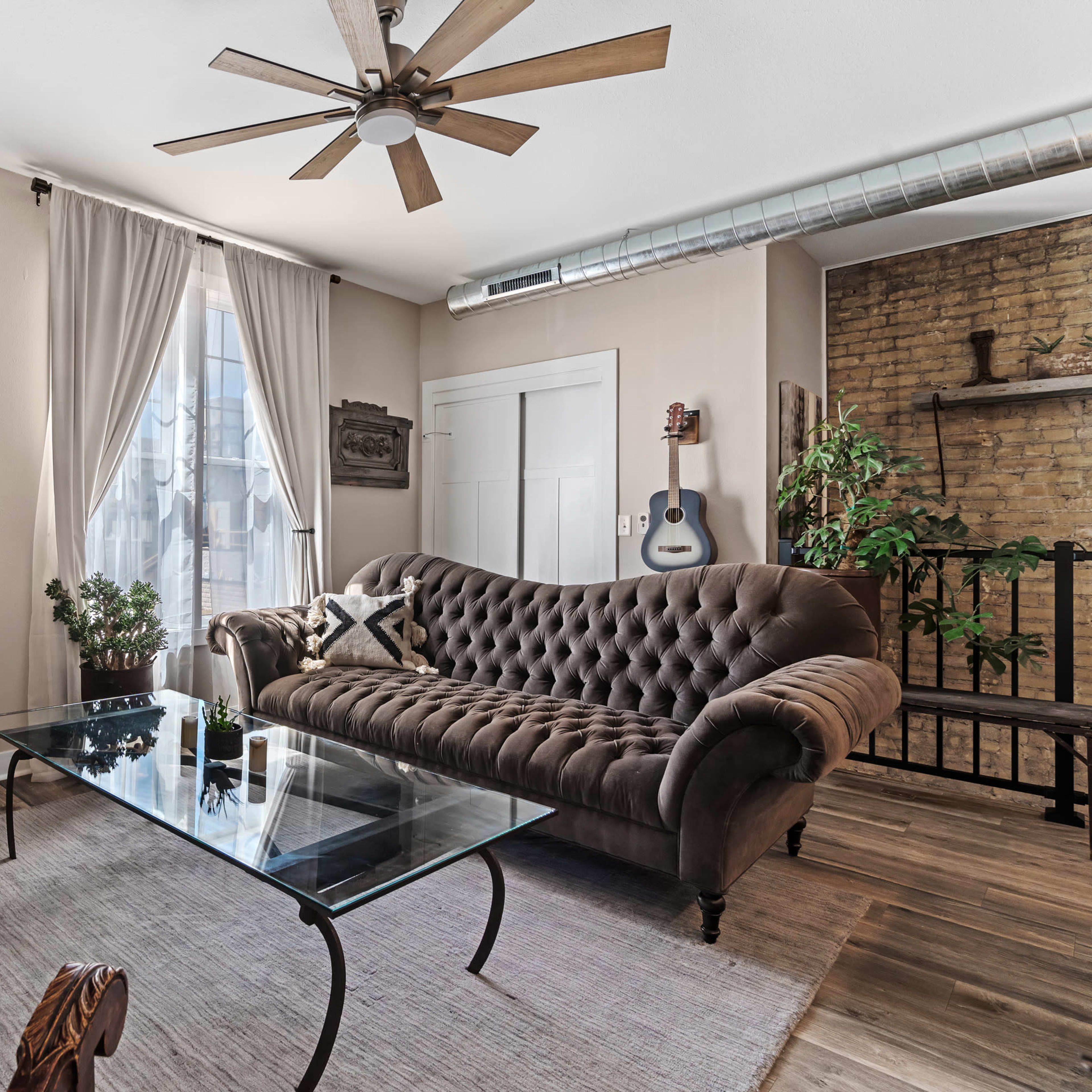 A living room featuring a brown tufted sofa, a glass coffee table, and a mix of natural plants, with a fan overhead and a guitar leaning against the wall.