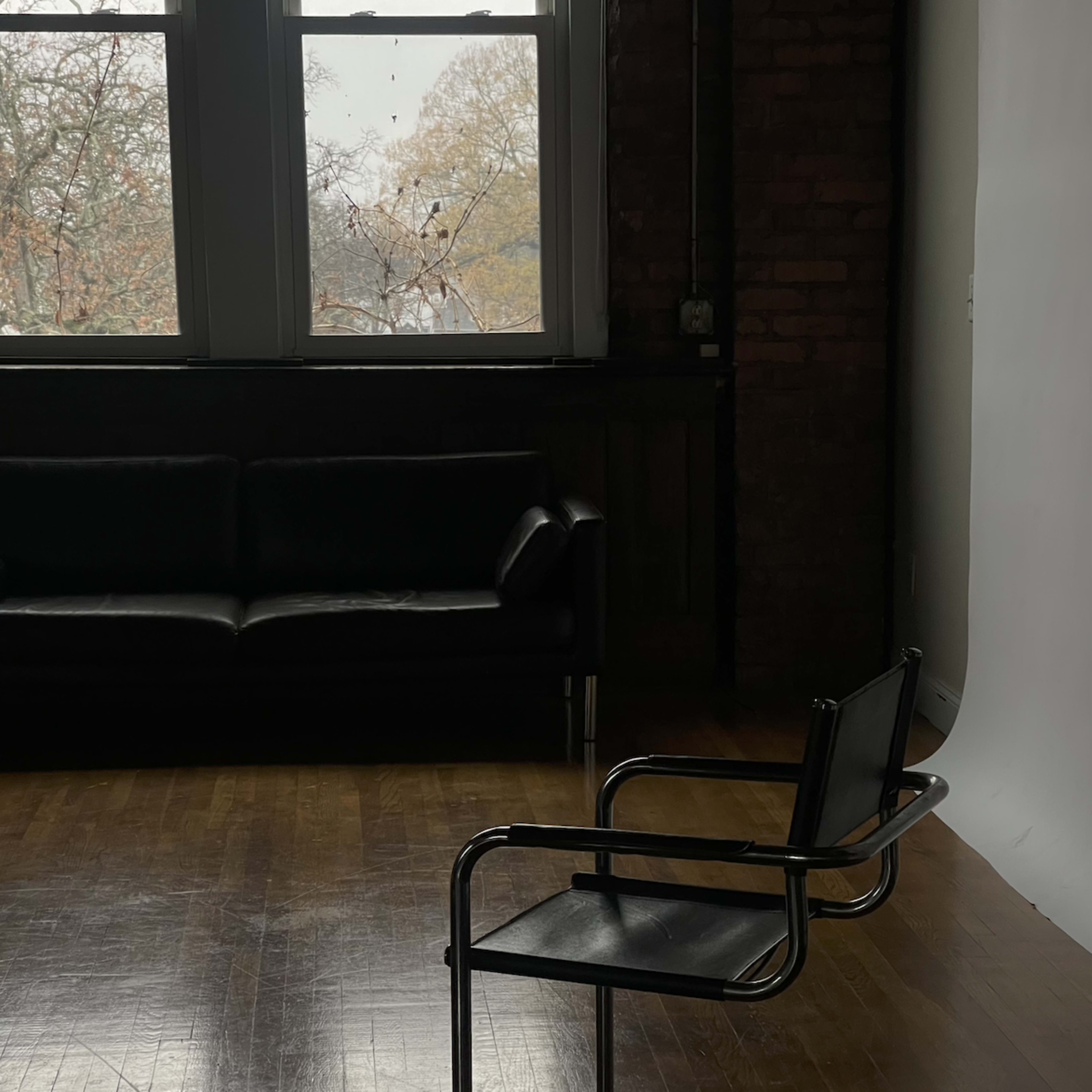 A metal-framed chair sits alone on a wooden floor with a dark couch and large windows in the background.