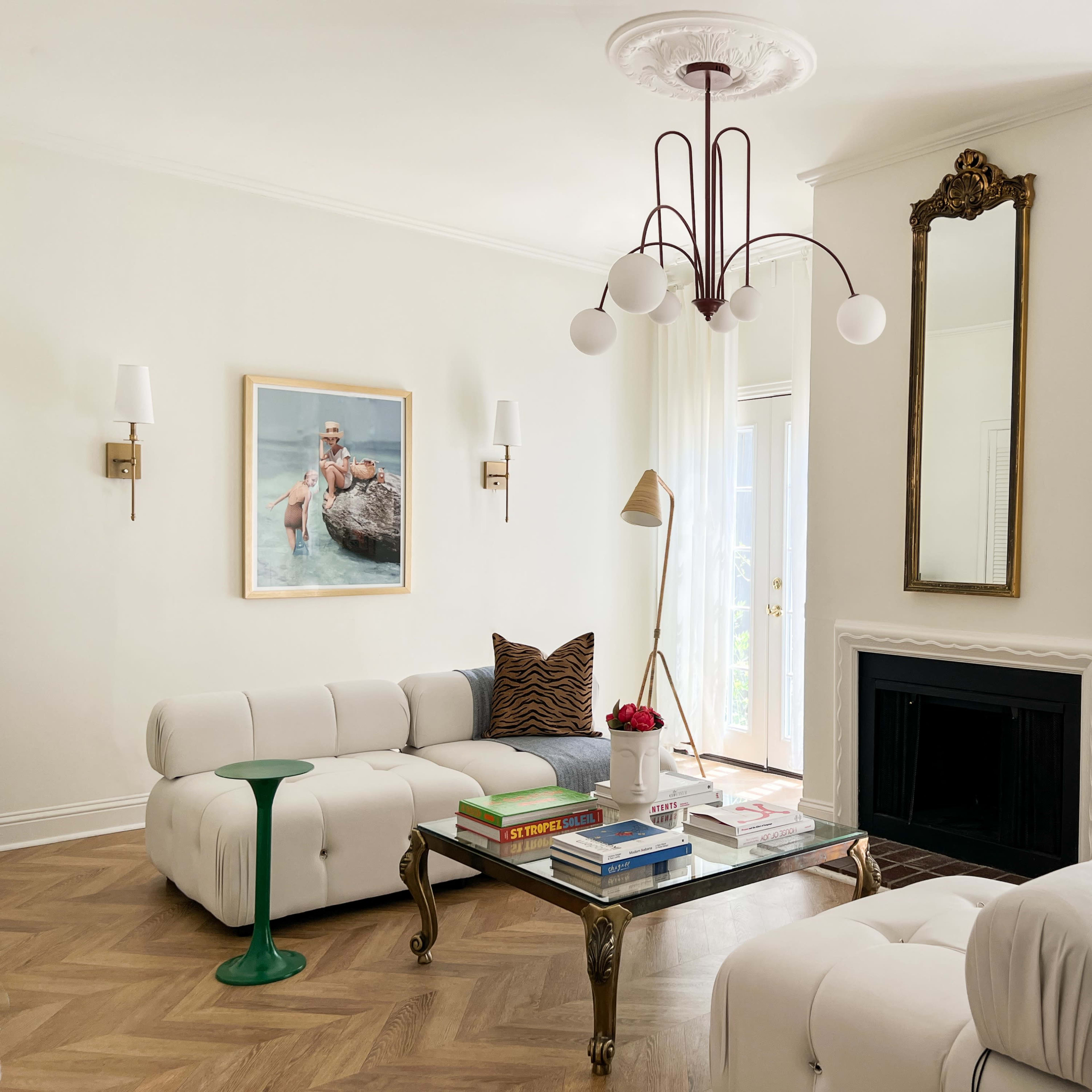 The living room features a light-colored sofa, a coffee table with books, a decorative mirror, and a large artwork on the wall, with floor-to-ceiling curtains and a fireplace.