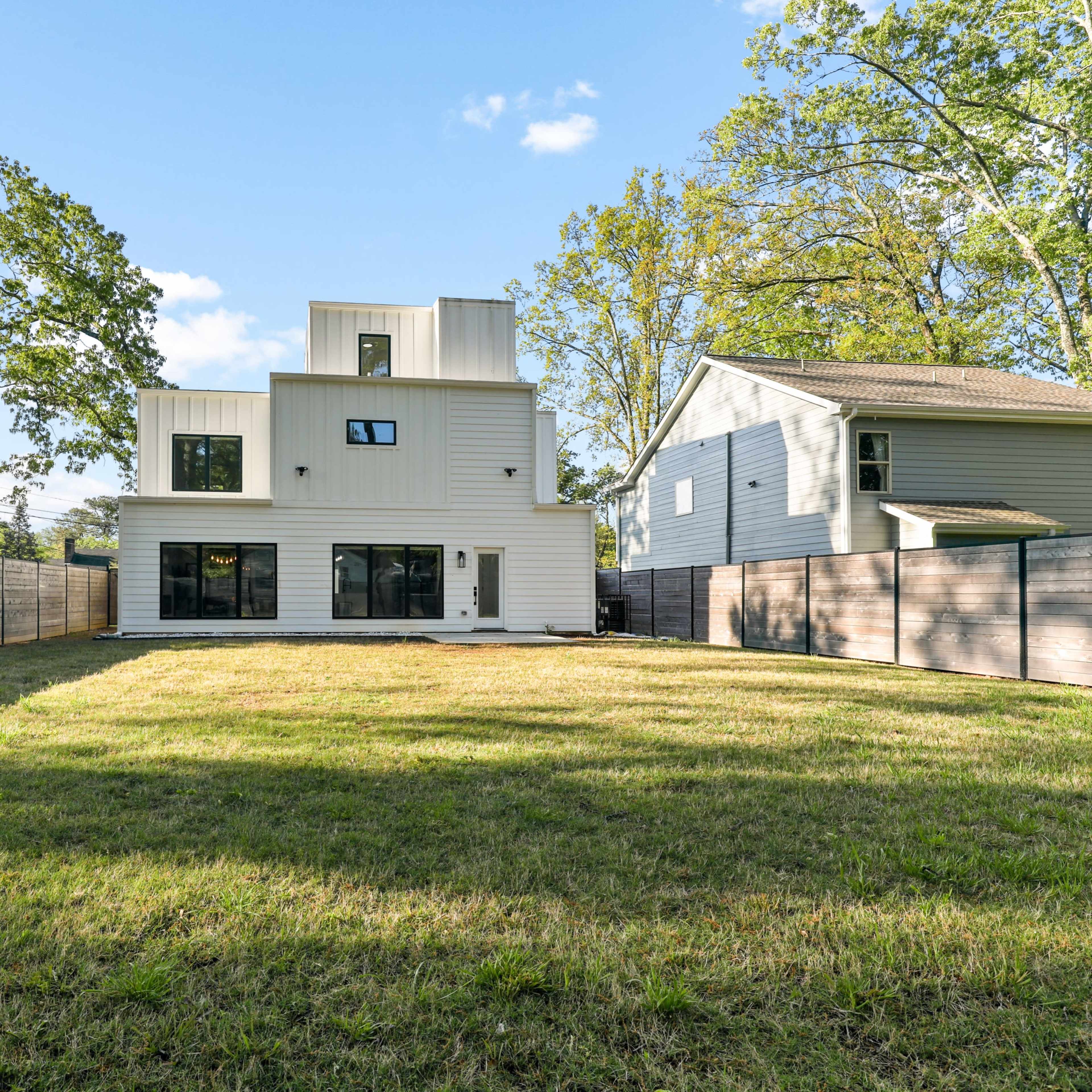 A modern two-story house with large windows, set in a spacious backyard enclosed by a wooden fence, with trees in the background.