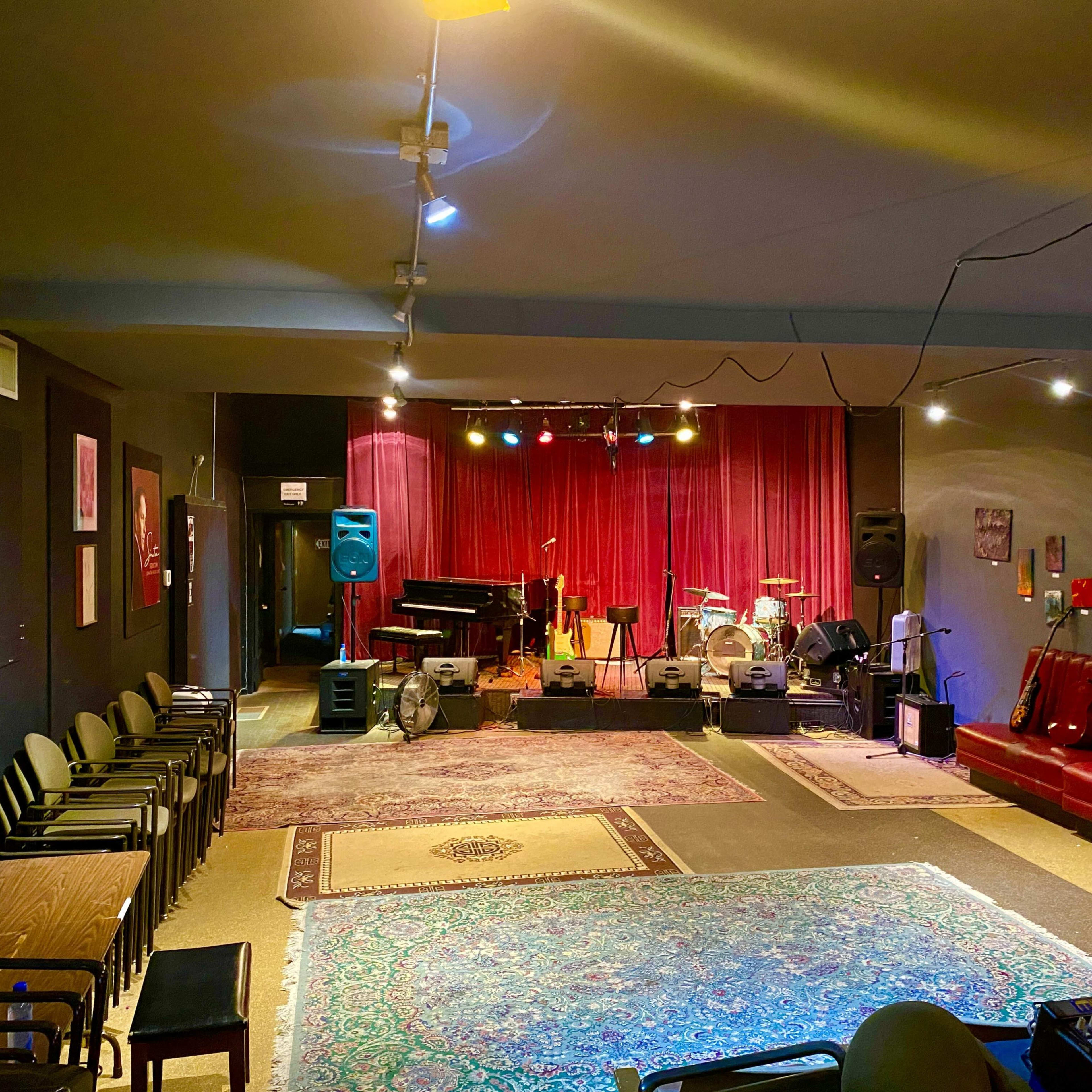 An empty music venue with a stage, red curtains, instruments, and seating arranged around the room.