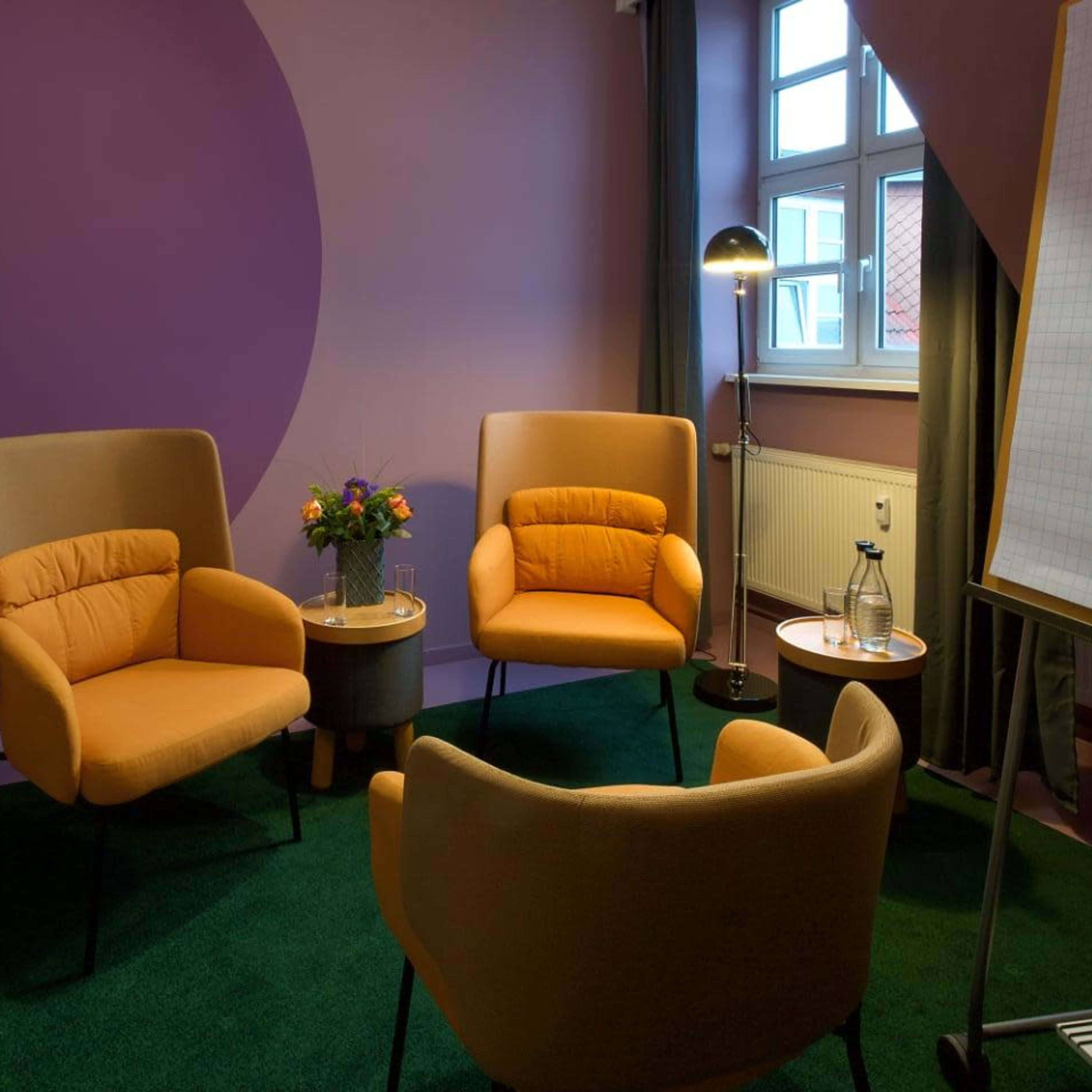 A modern meeting room with three orange chairs, a small round table, a lamp, and a whiteboard against a purple and pink wall.