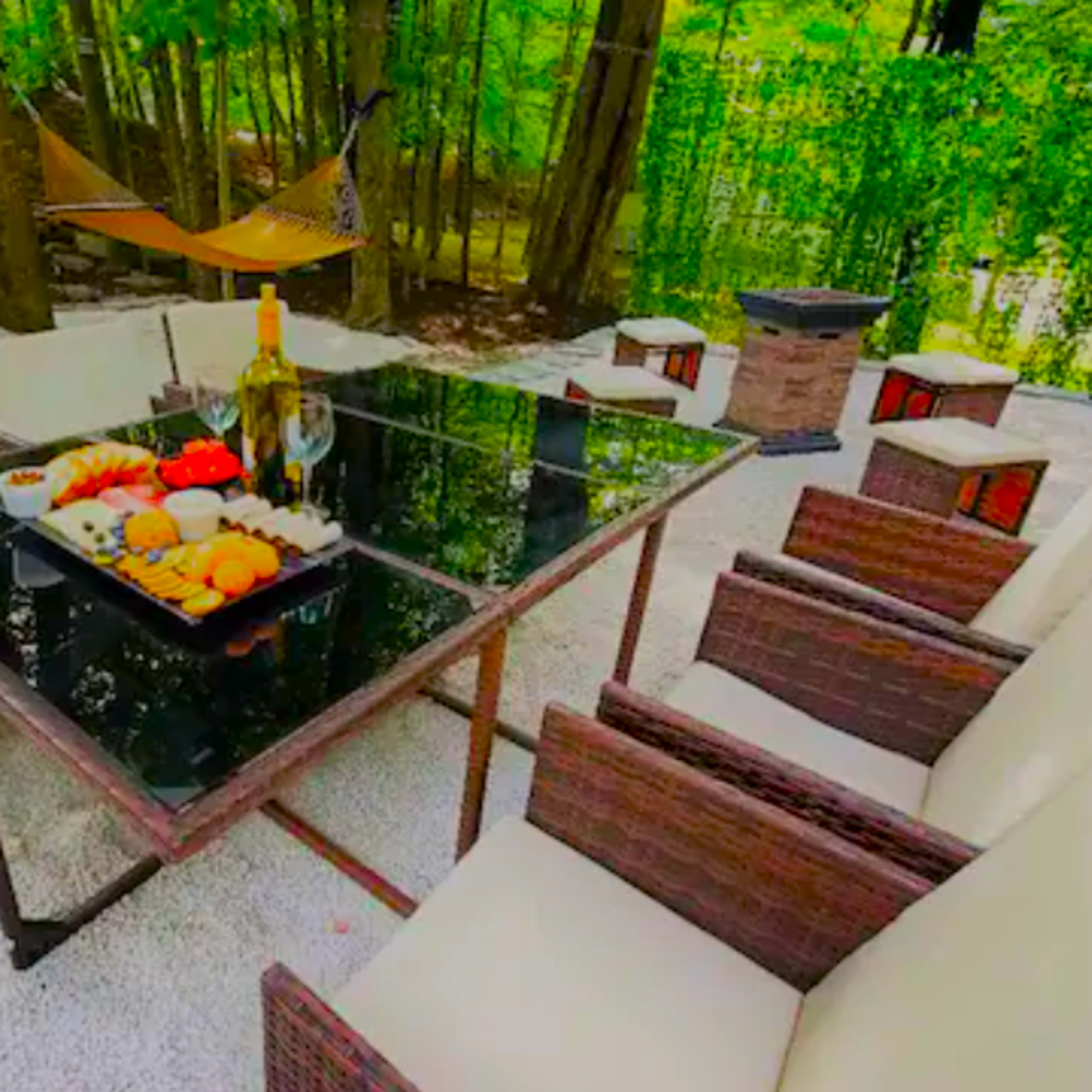 A dining table with a tray of fruits and a bottle of wine is surrounded by several chairs in a wooded outdoor setting.