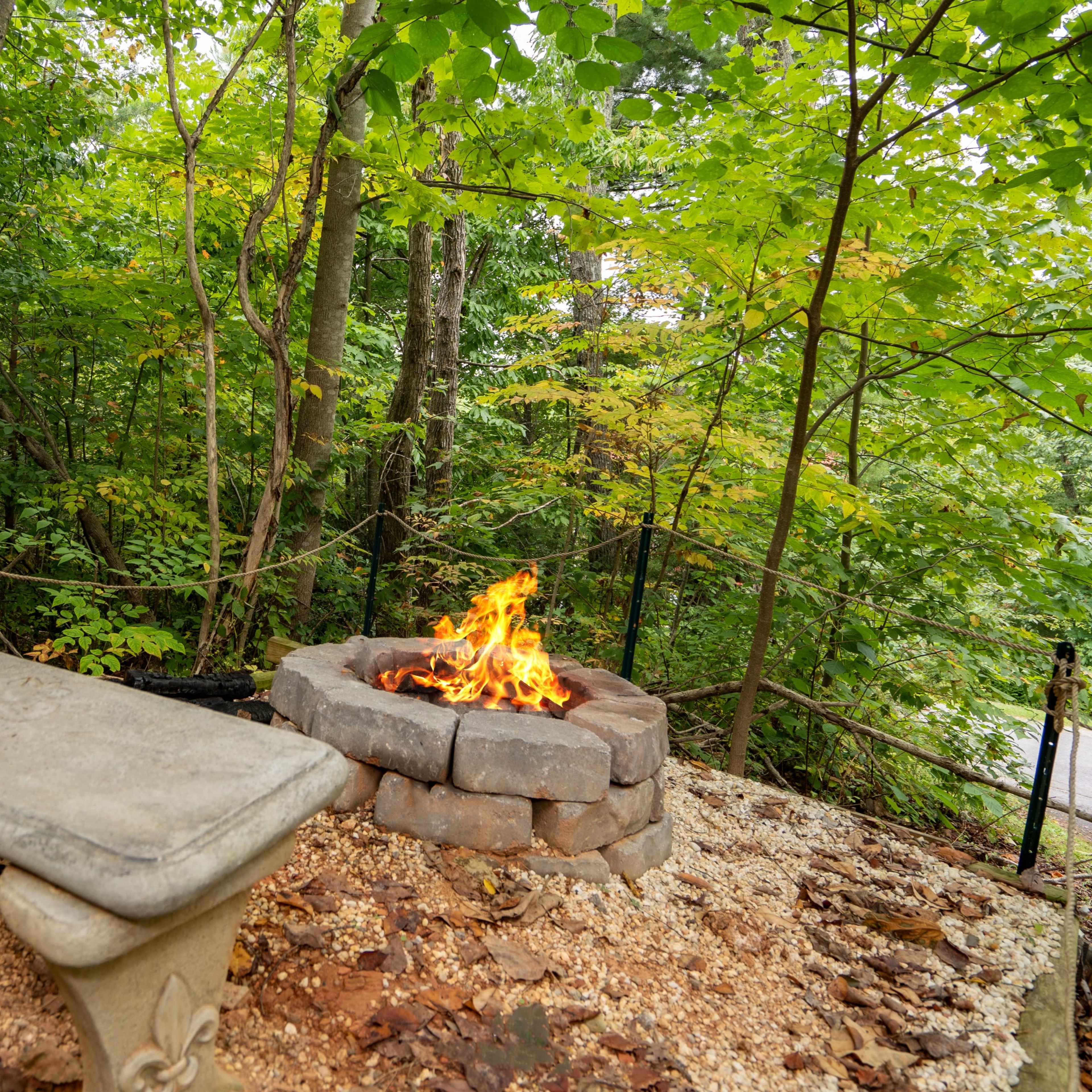 A stone fire pit surrounded by a concrete bench is nestled in a wooded area with green trees and fallen leaves.