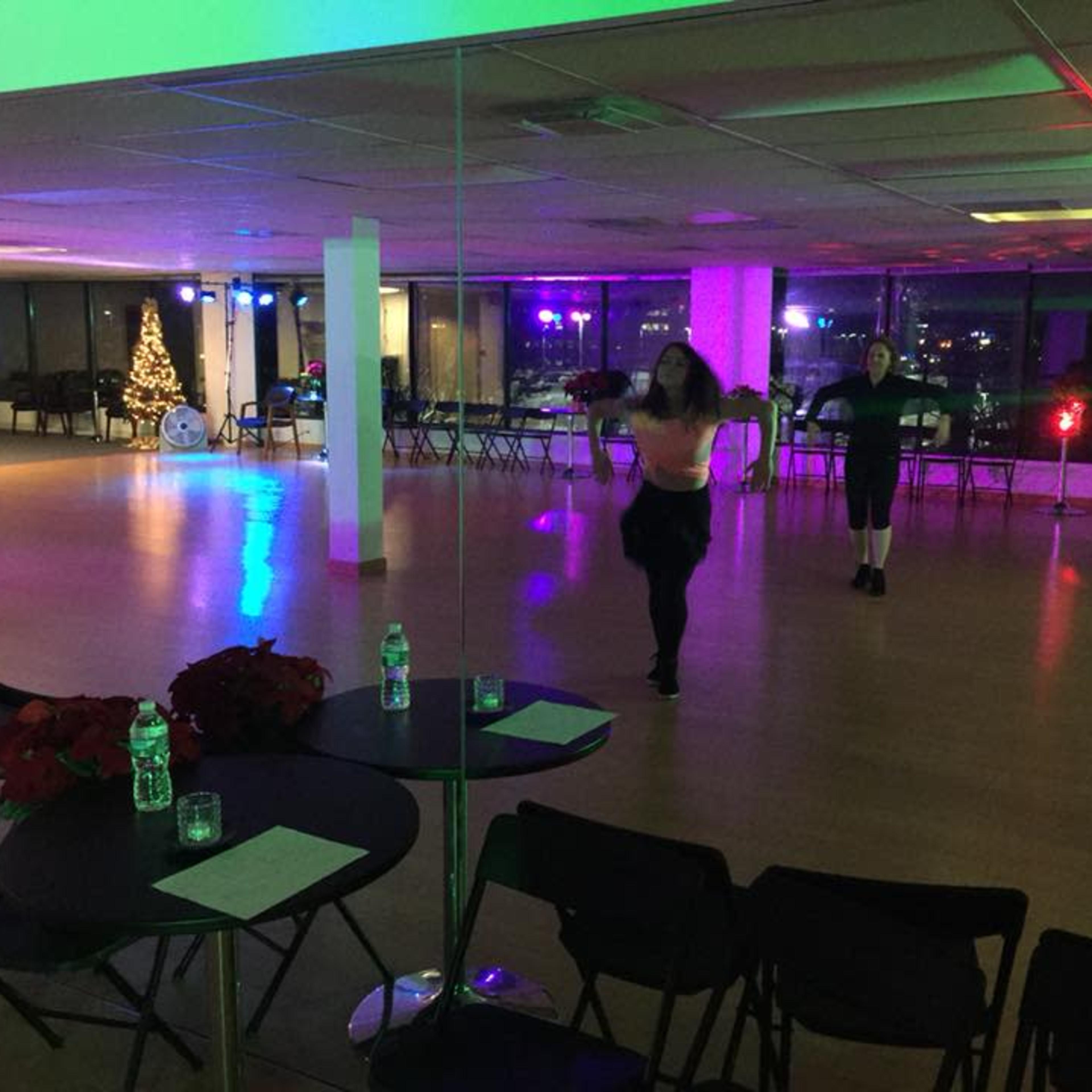 Two dancers practice in a spacious dance studio with mirrored walls and festive decorations, including a Christmas tree, under colorful lights.