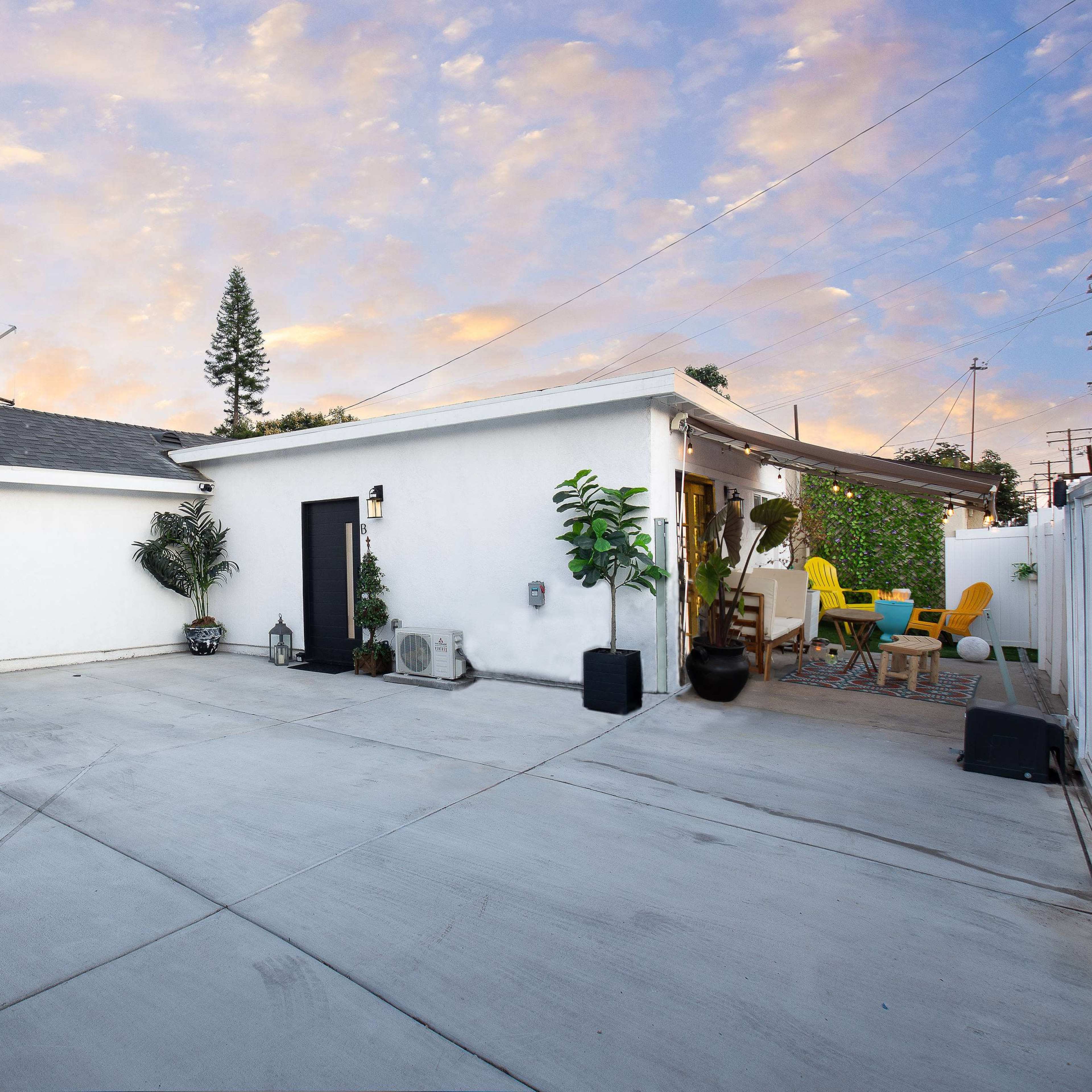 A concrete backyard with a white stucco building, a black storage shed, and a seating area surrounded by greenery.