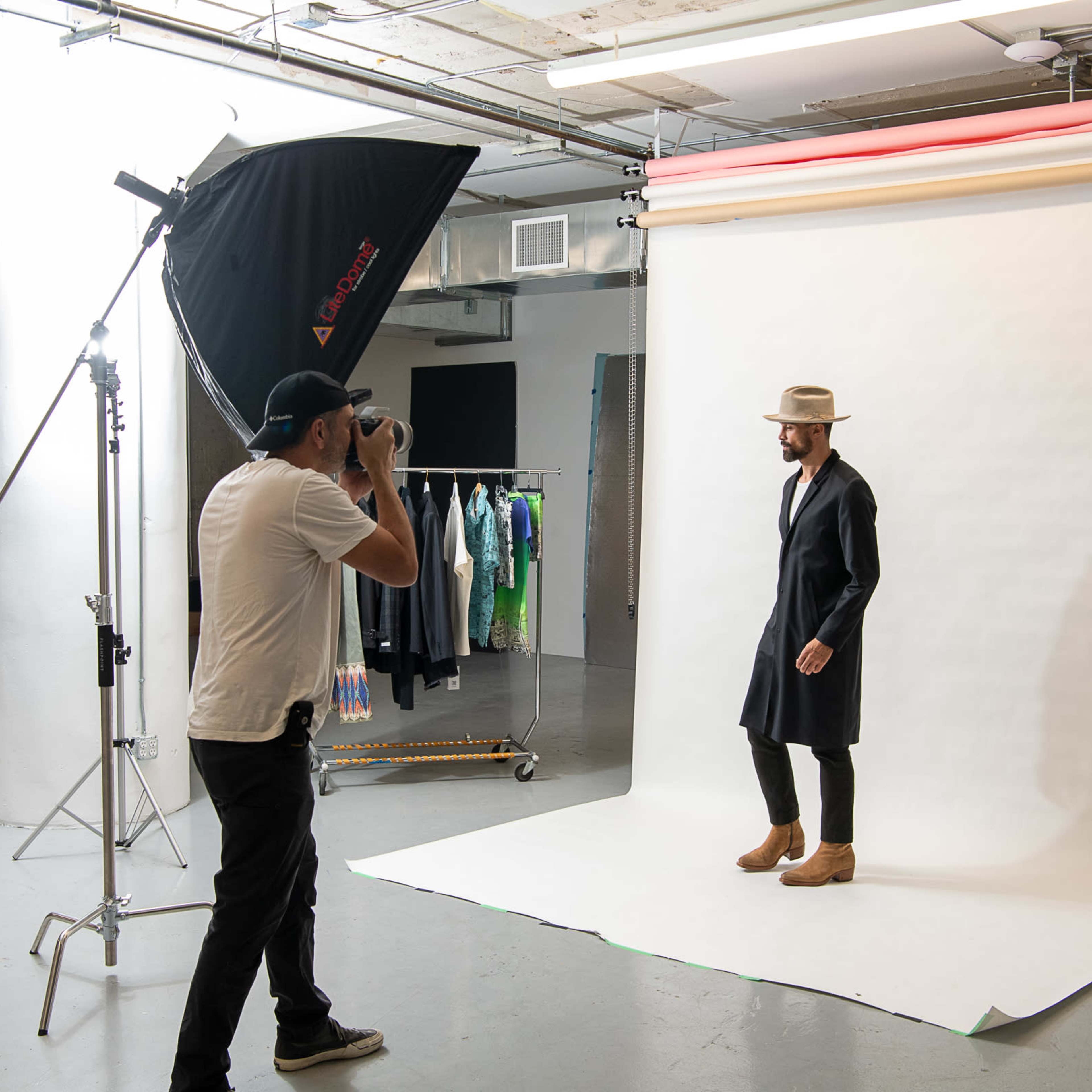 A photographer captures a man posing in a long coat and hat against a plain white backdrop in a studio.