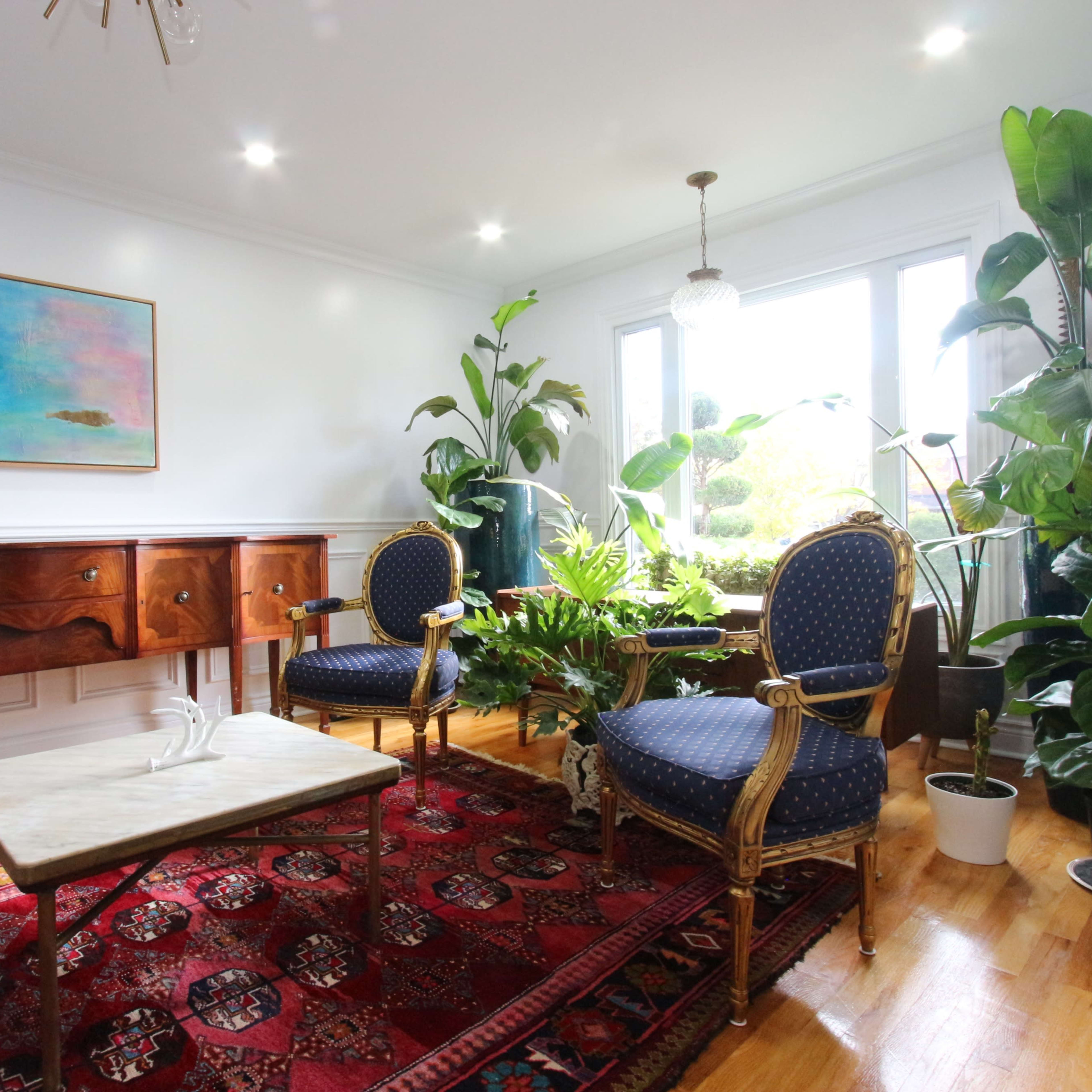 A well-lit living room with two blue upholstered chairs, a marble-top coffee table, a wooden sideboard, and several potted plants, all set against a colorful wall art piece.