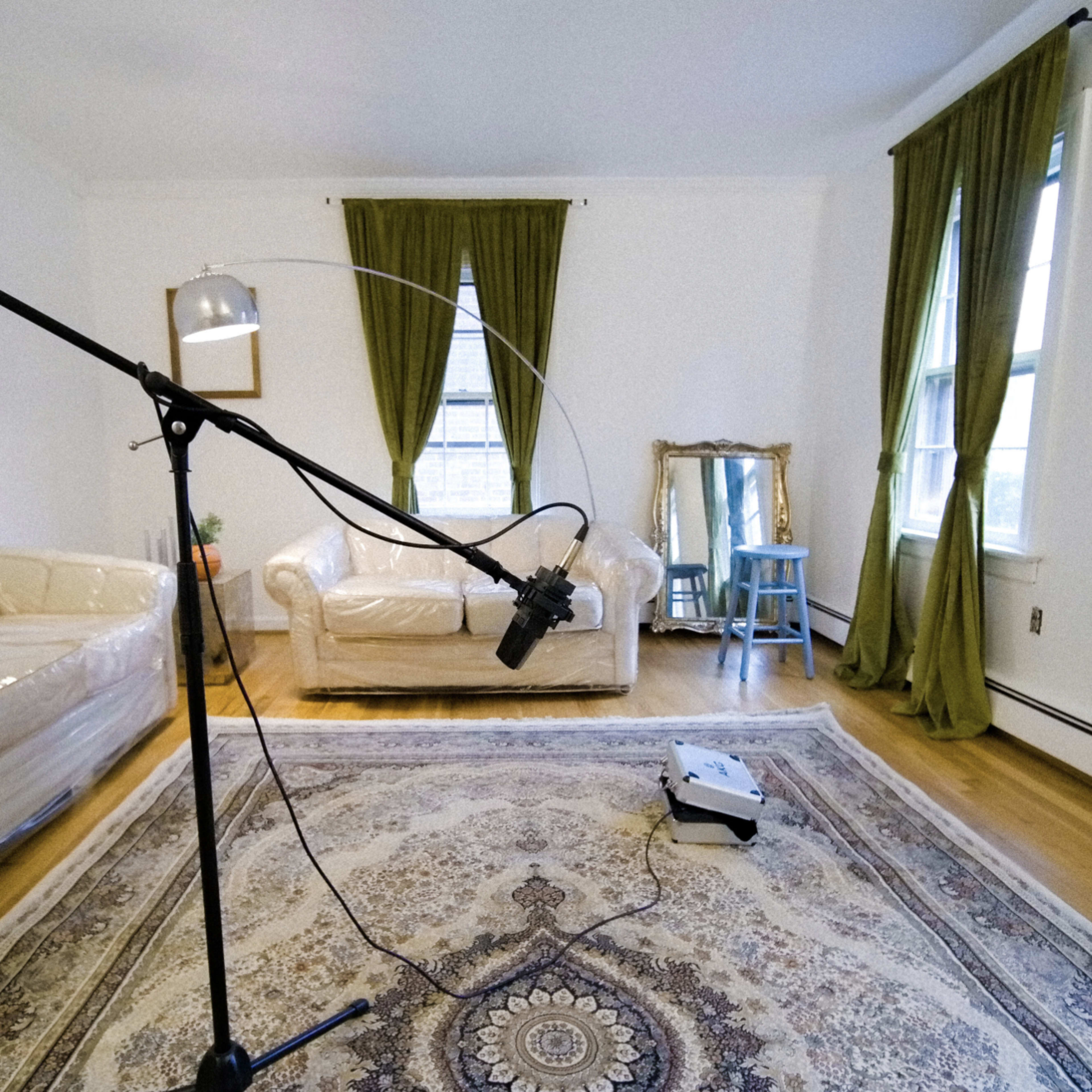 A living room with a clear plastic-covered couch, a microphone setup on a stand, a large area rug, and green curtains at the windows.