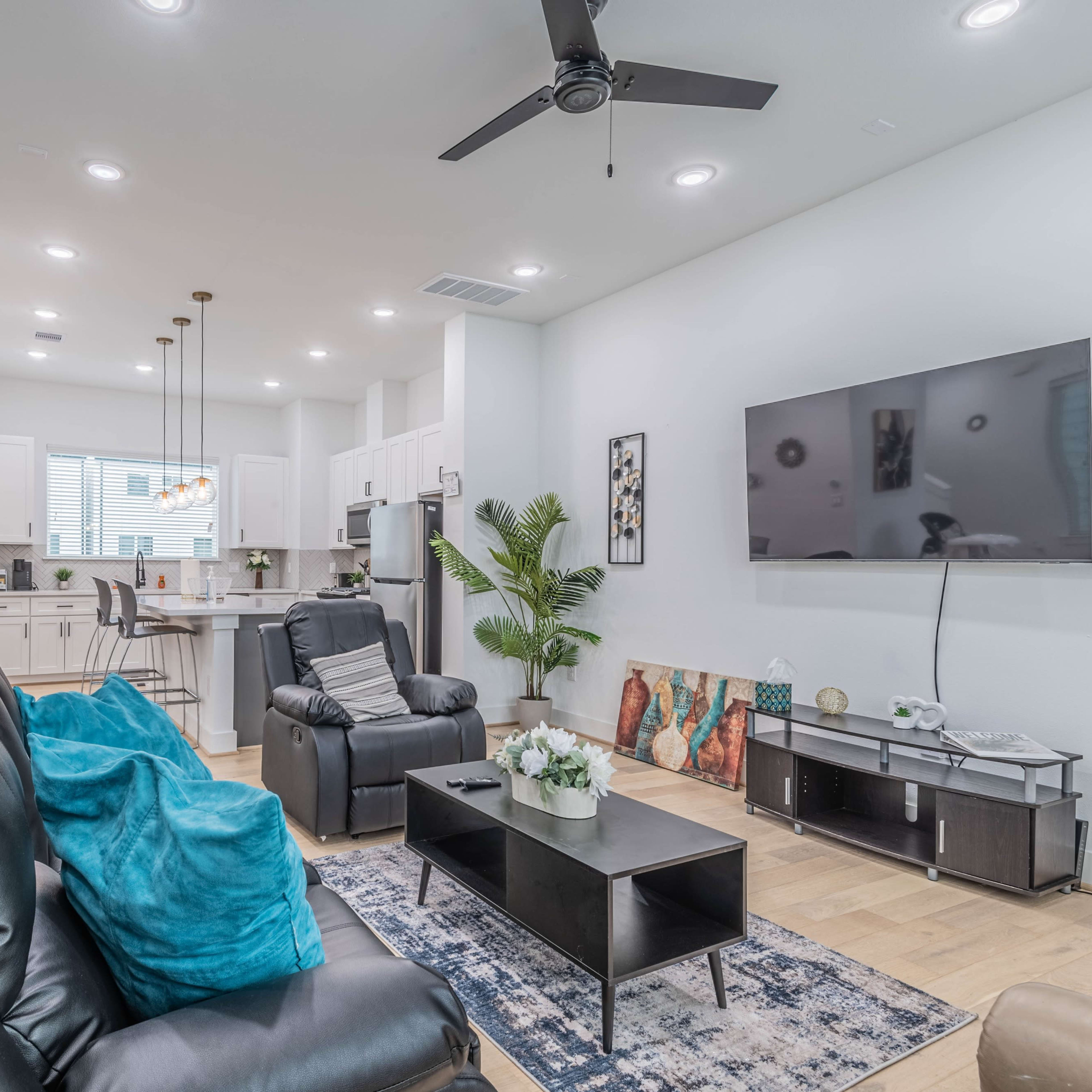 A modern living room with black recliners, a television mounted on the wall, a coffee table, and a kitchen area visible in the background.