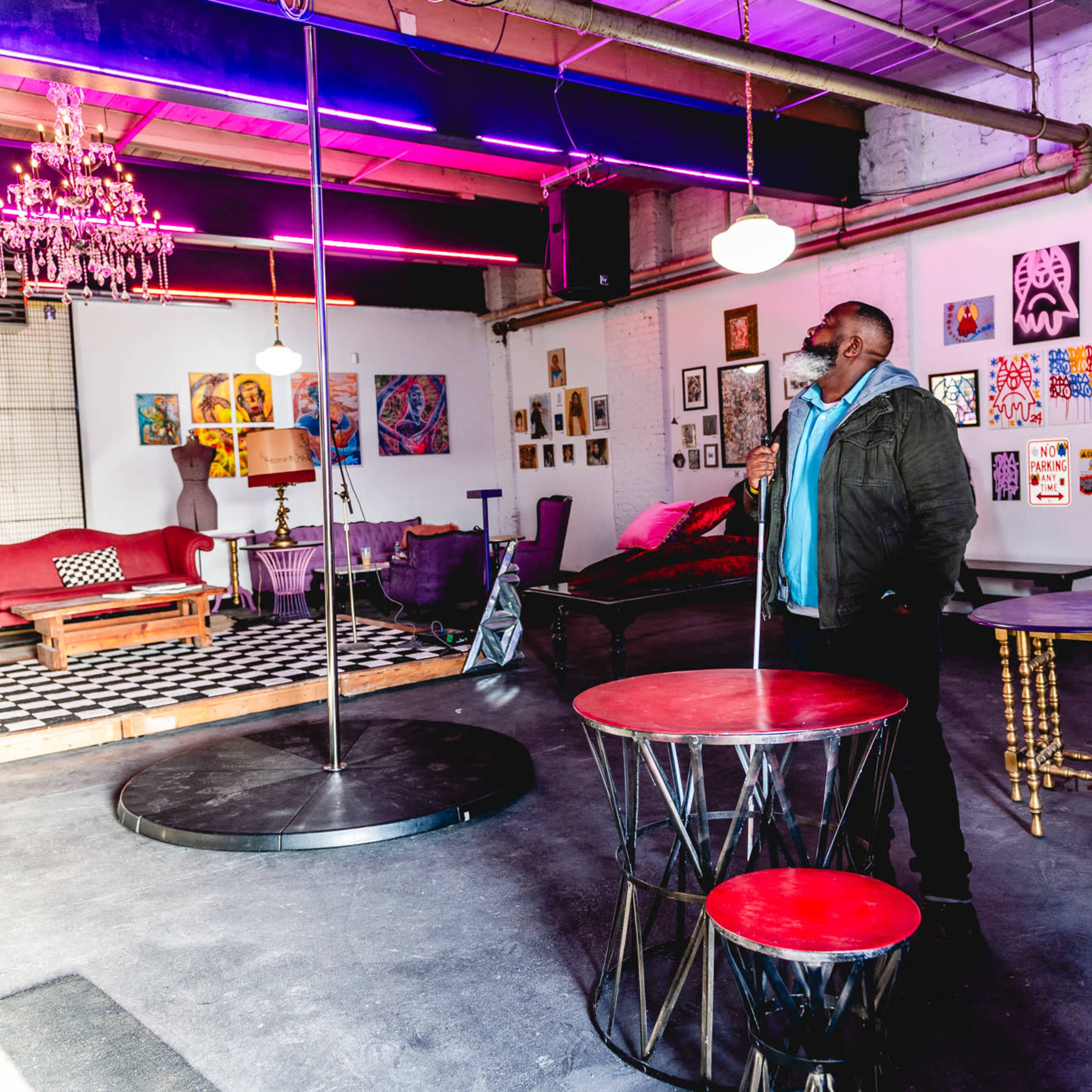 A man stands observing the interior of a brightly lit art studio featuring a dance pole, eclectic furniture, and various artworks displayed on the walls.