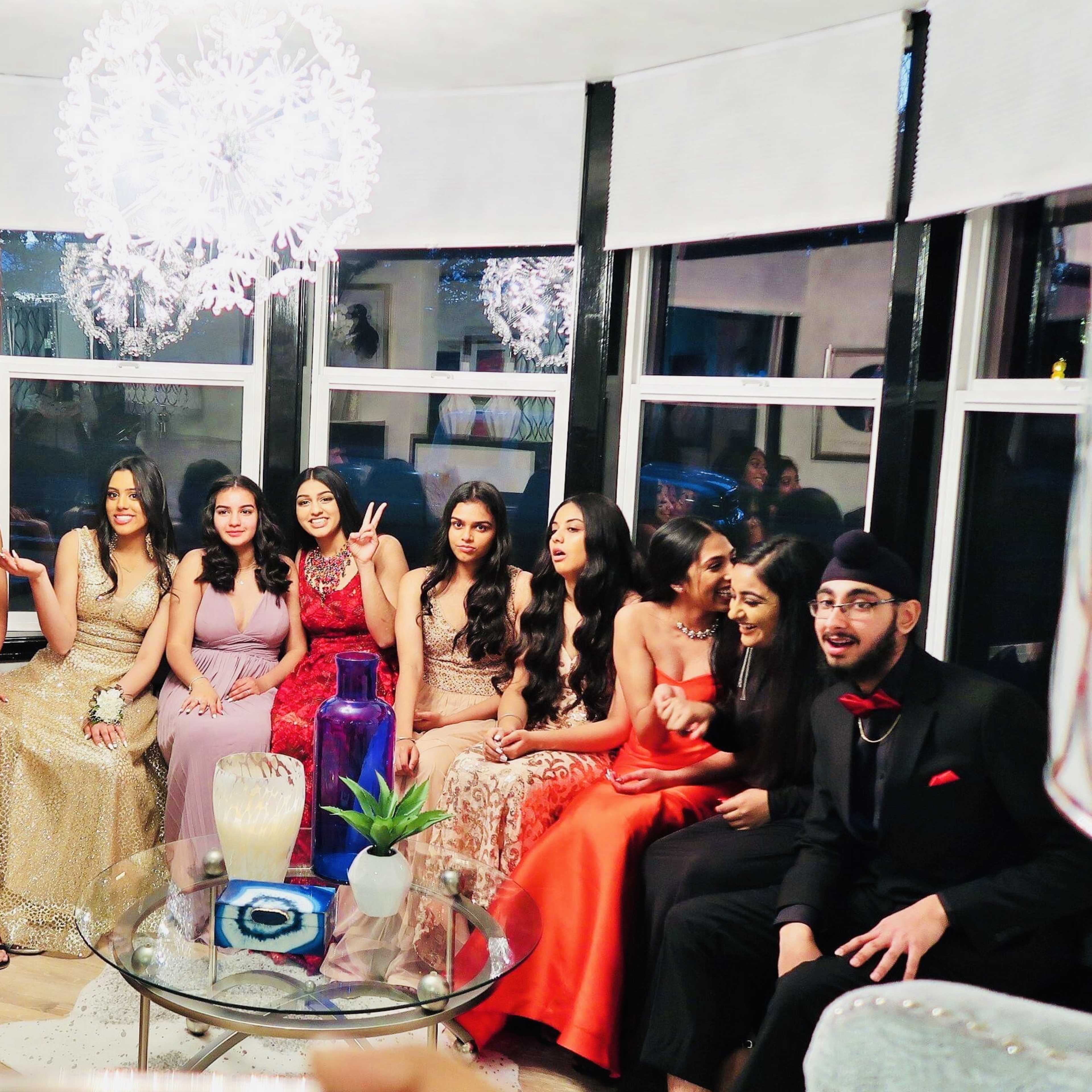 A group of thirteen young adults in formal attire poses together in a decorated living room with large windows and modern lighting.