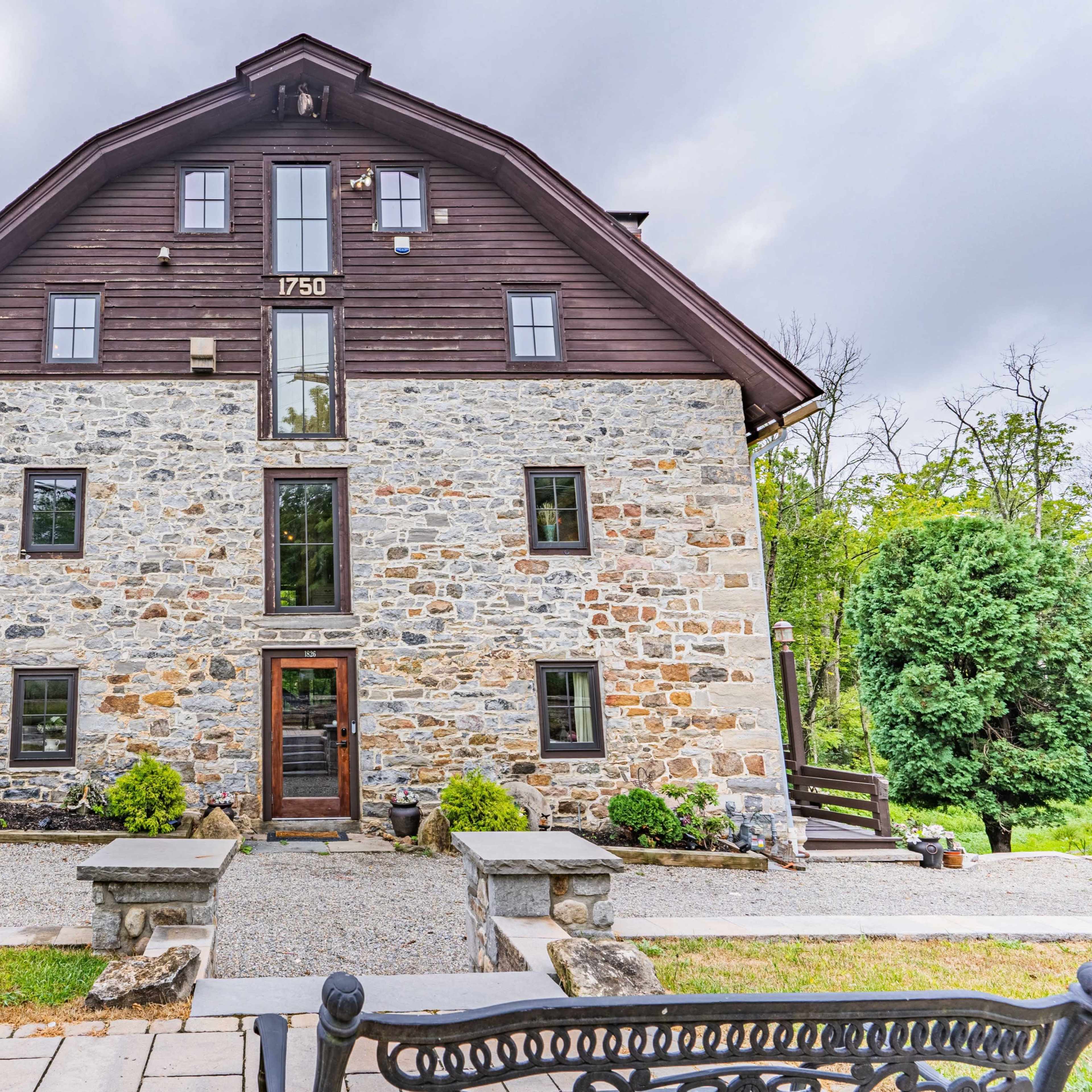 A three-story stone building with a triangular roof has a wooden upper section, set in a landscaped area with a gravel pathway.