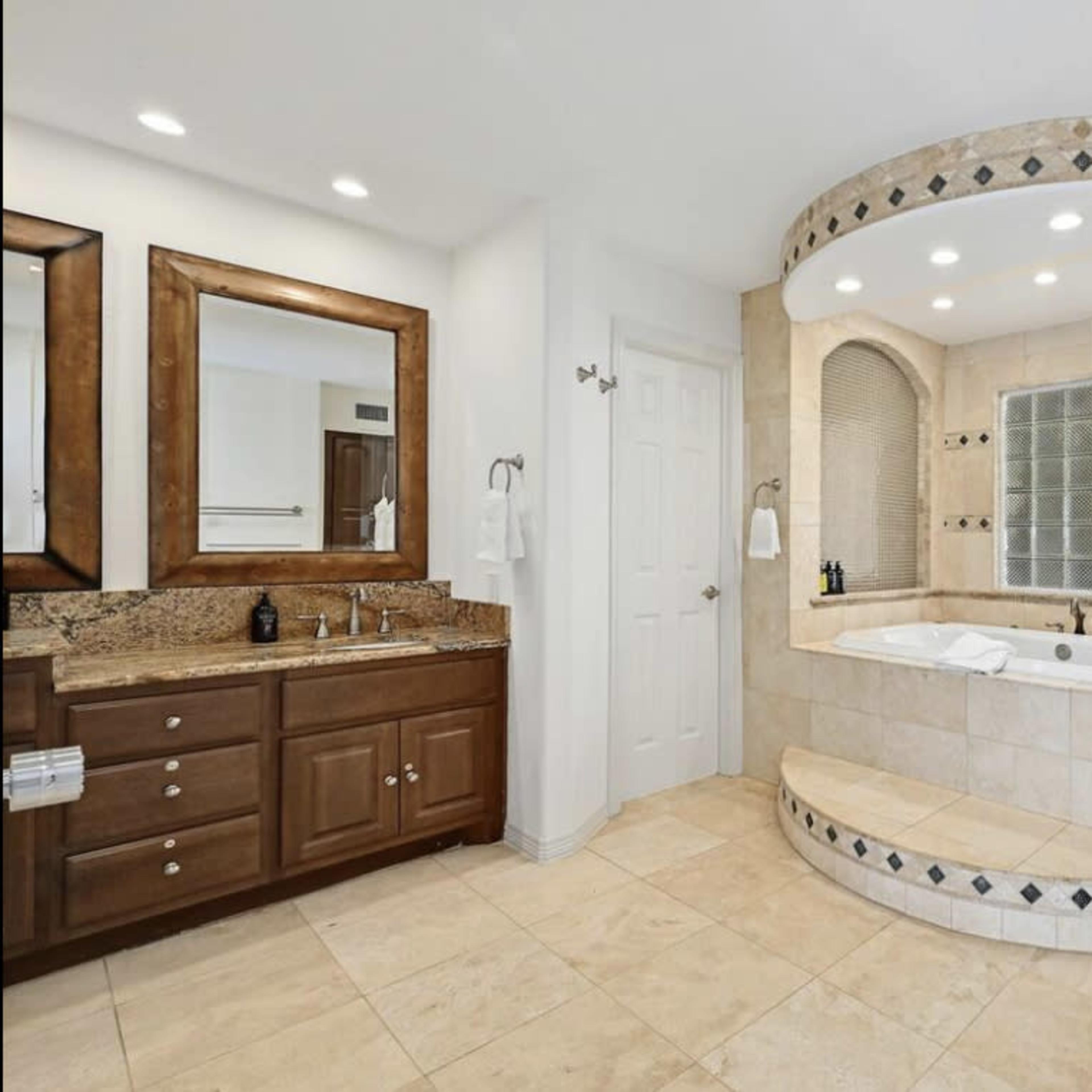 A spacious bathroom featuring a large tub with steps, a glass block window, a walk-in shower, and double vanities with mirrors.