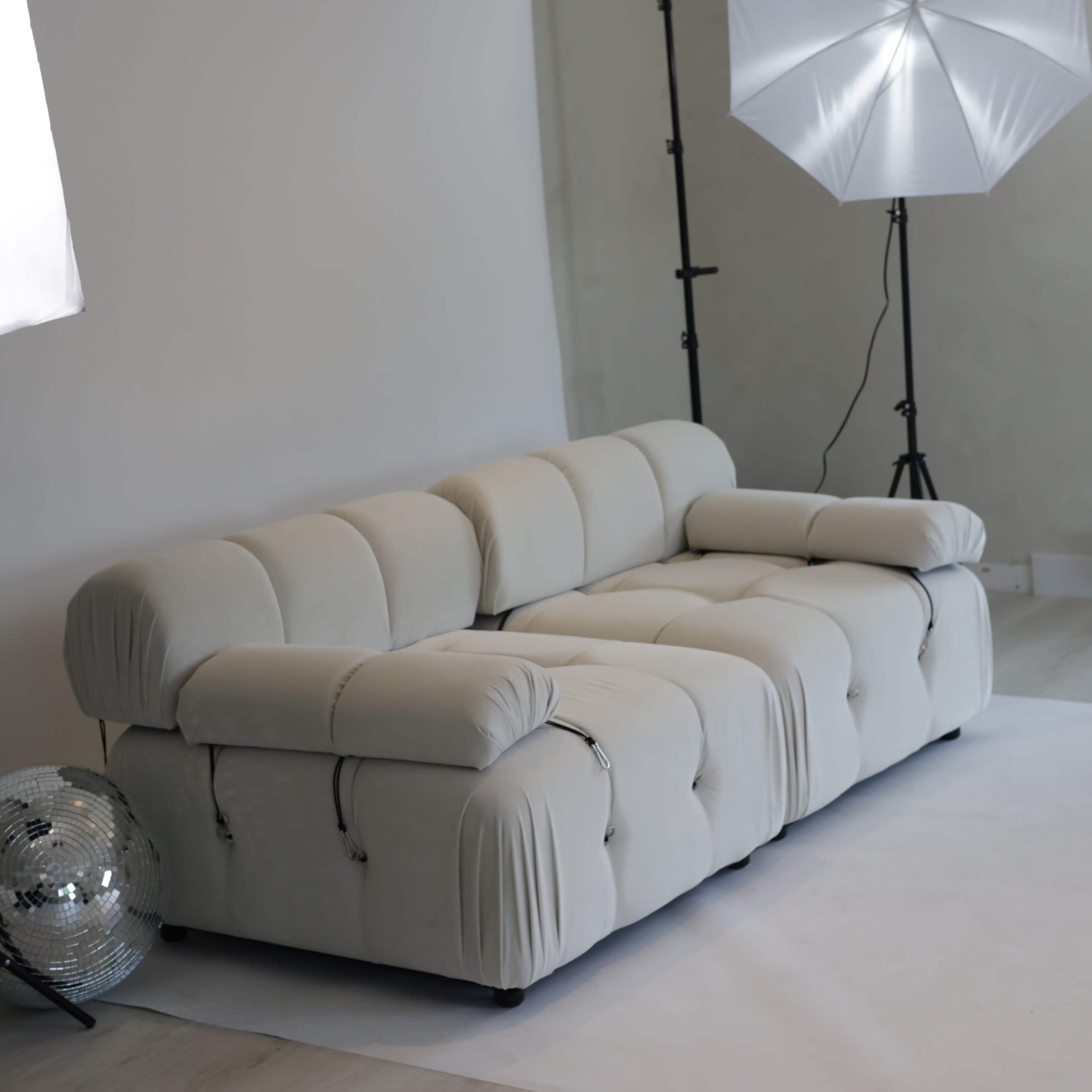 A beige sectional sofa is positioned in a minimalistic studio setting, accompanied by two softbox lights and a mirrored disco ball.