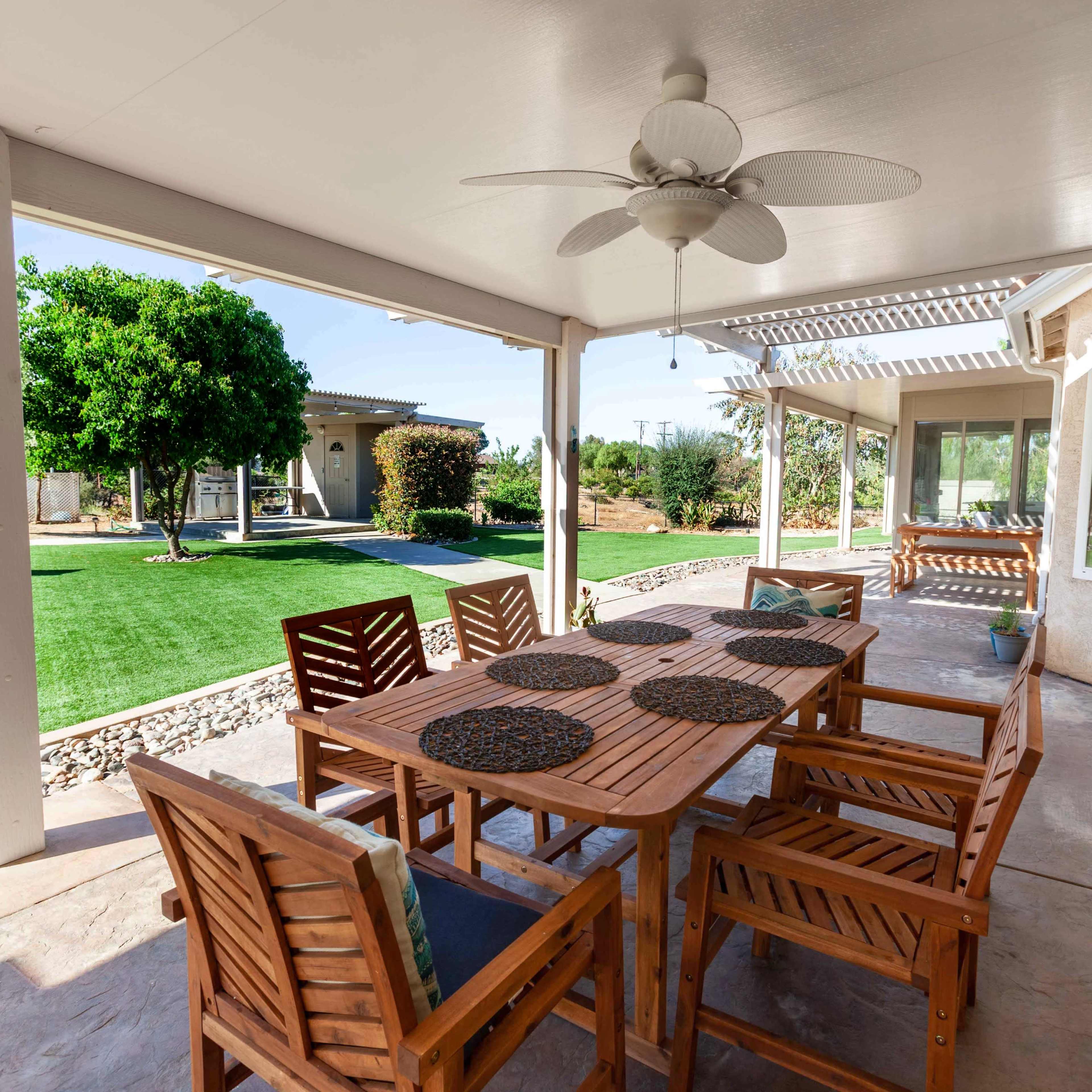 A wooden dining set is arranged under a covered patio, overlooking a lawn and landscaped yard.