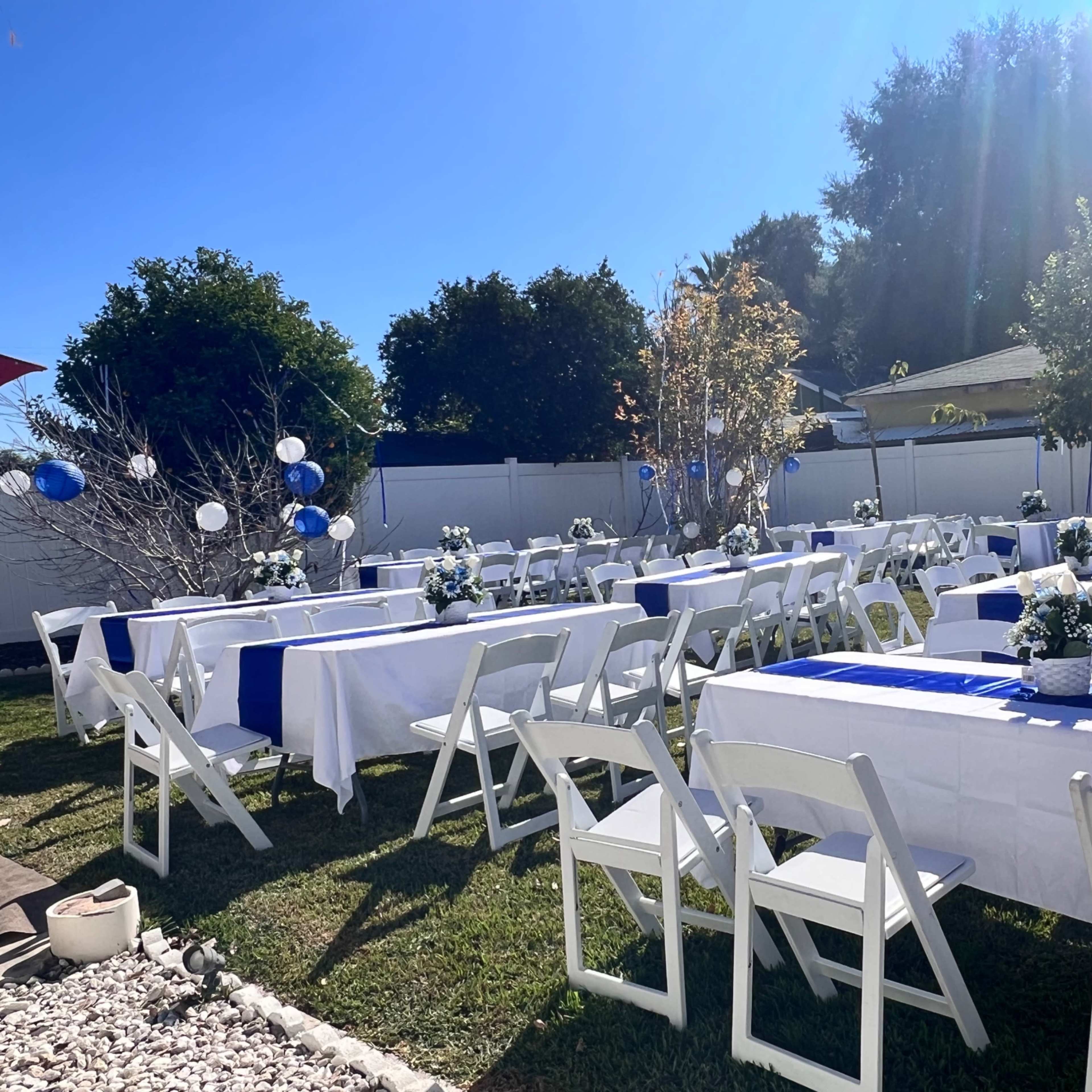 A neatly arranged outdoor event space with several white tables set with blue table runners and floral centerpieces, surrounded by green grass and trees.