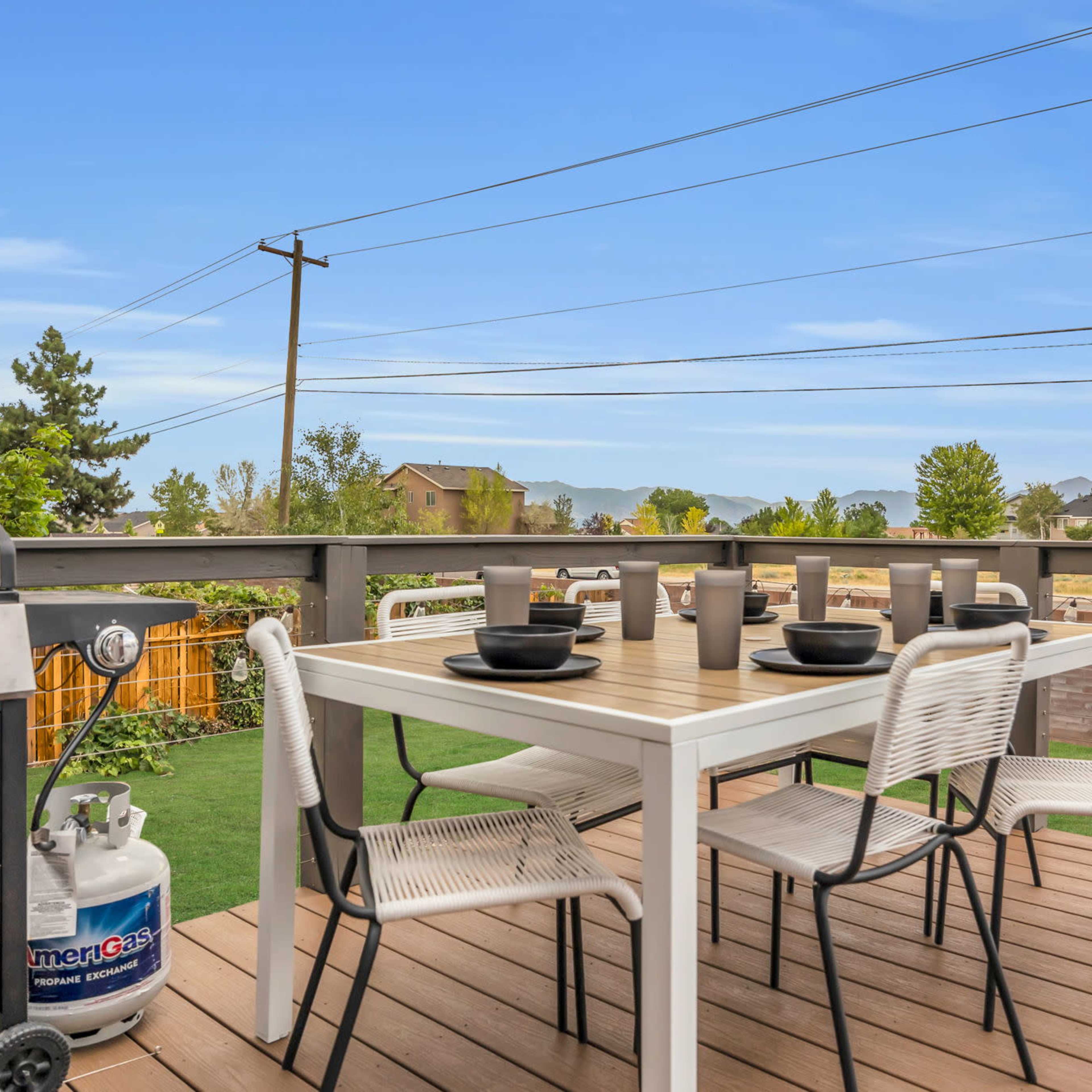 A wooden deck features a outdoor dining table set with plates and cups, accompanied by a grill and a propane tank in the foreground.