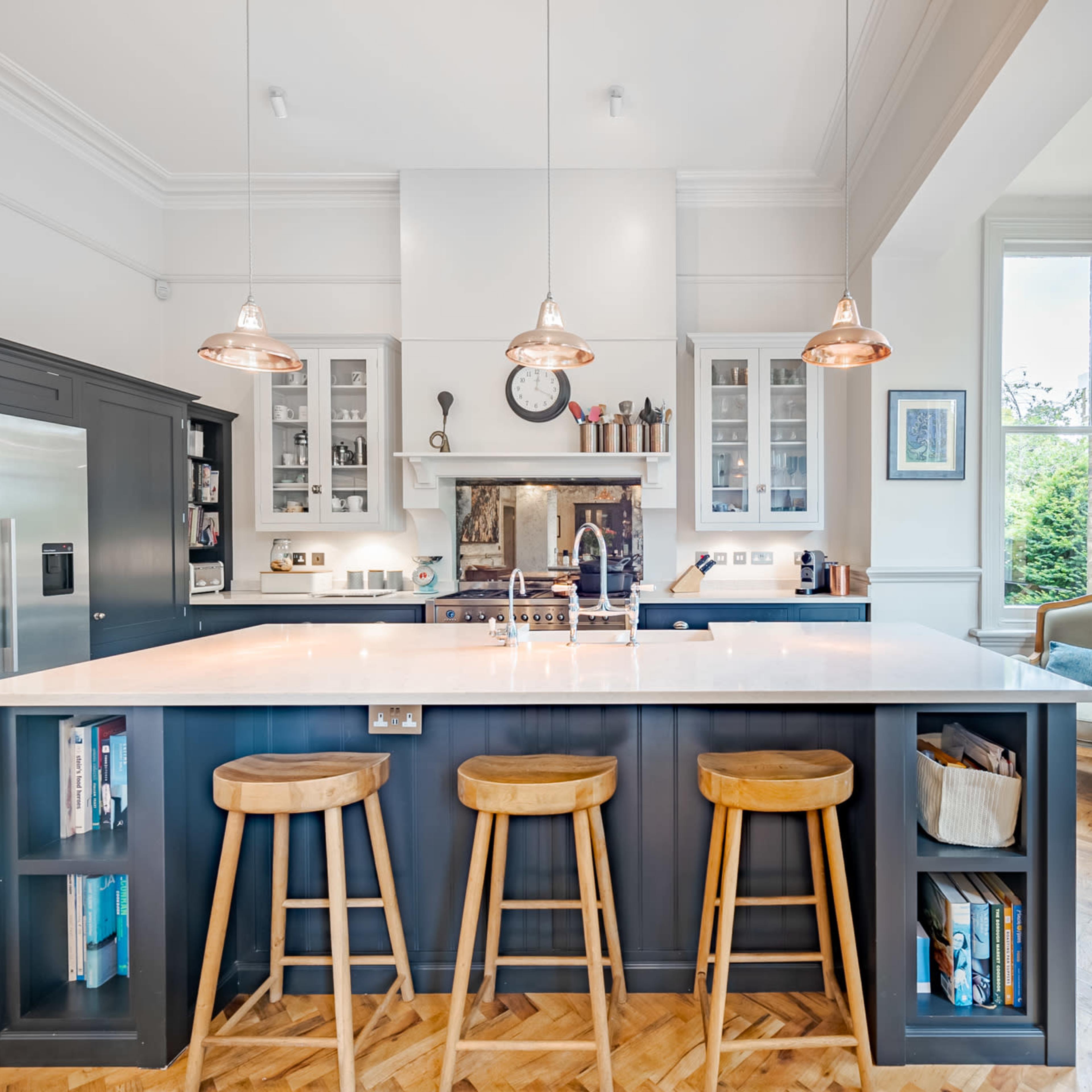 A modern kitchen with a large island, three wooden barstools, stainless steel appliances, and large windows allowing natural light to flow in.