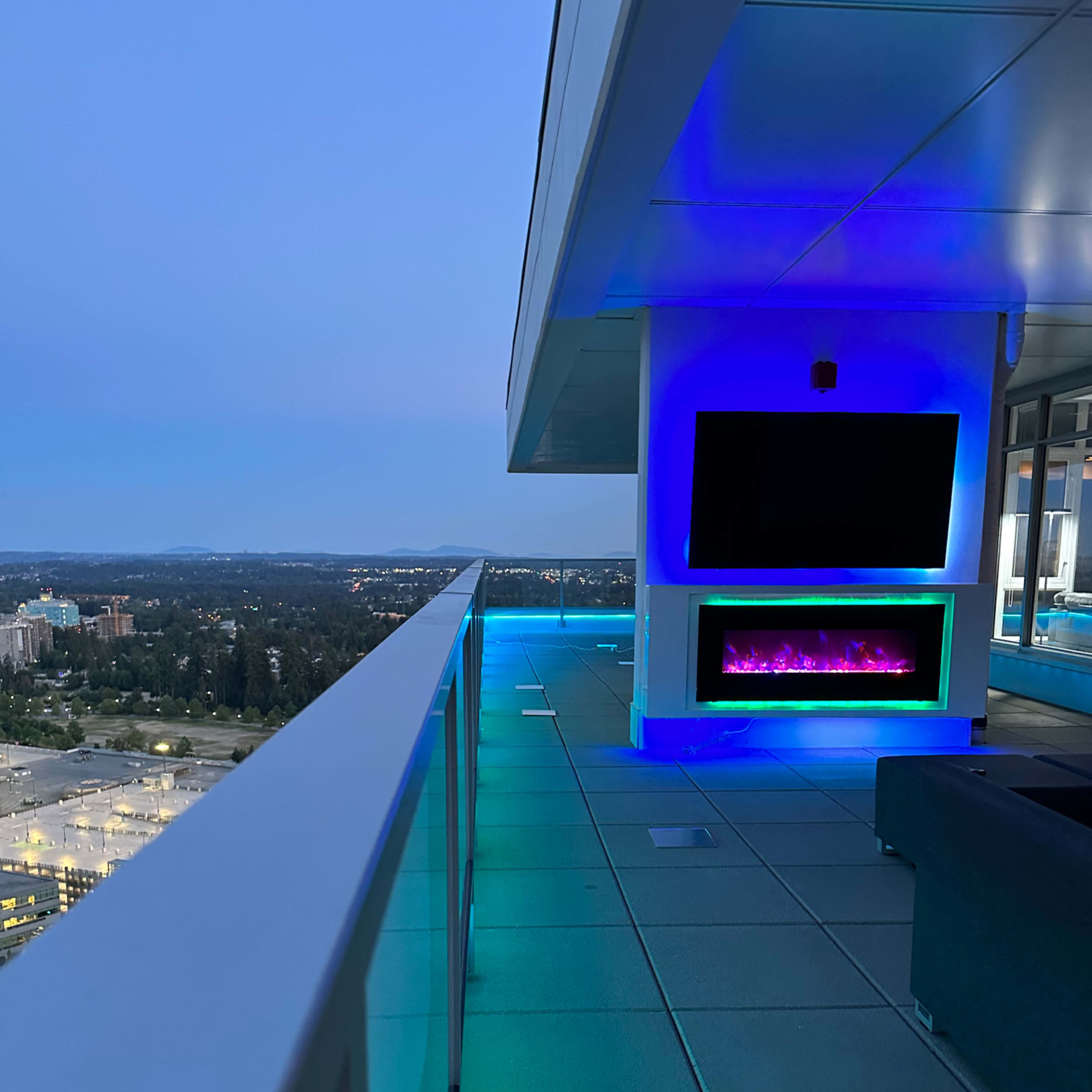 A balcony with modern lighting features, a television mounted on the wall, and a view of a city skyline at dusk.