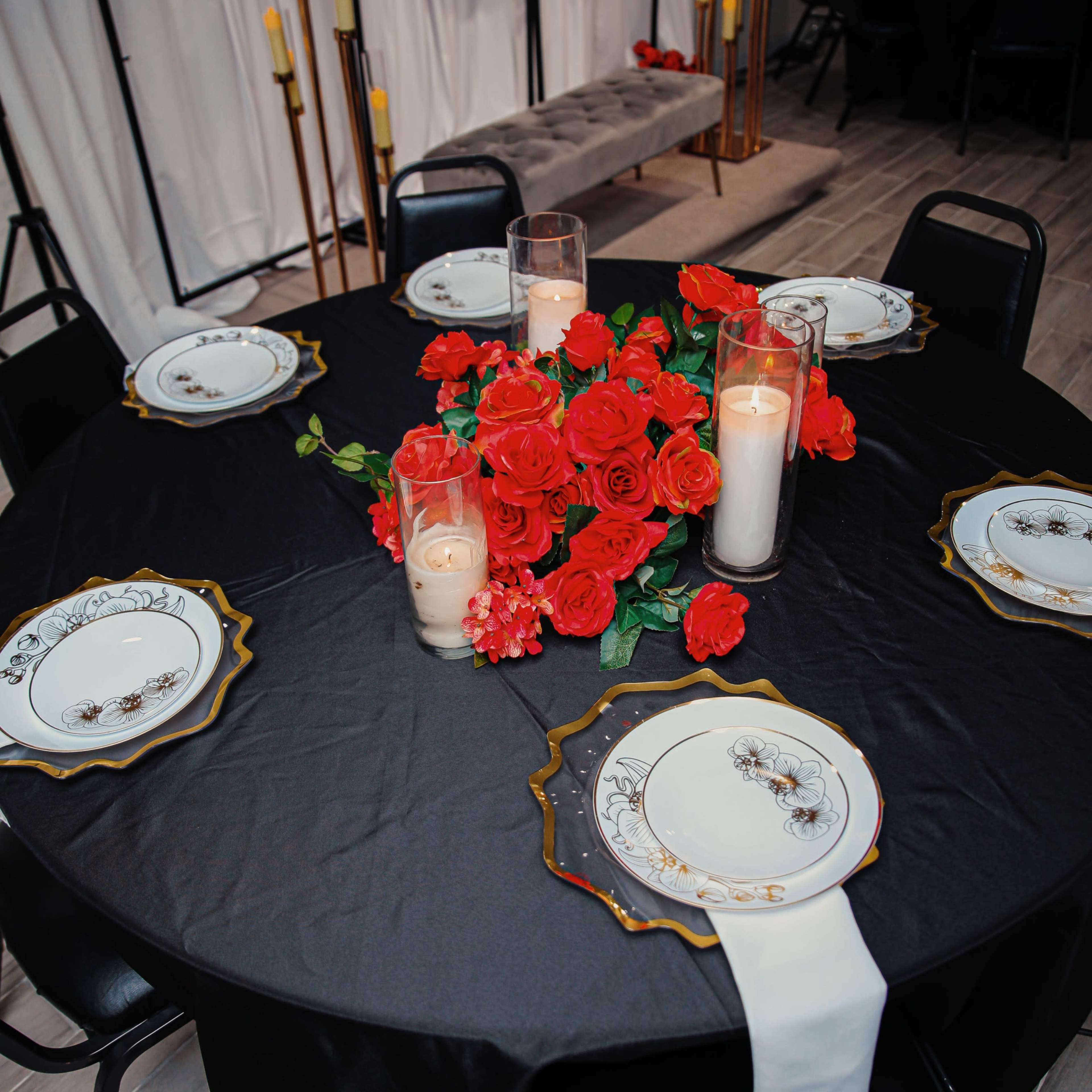 A round black table is set with white plates, red roses, and candles, creating a decorative centerpiece in a dimly lit room.