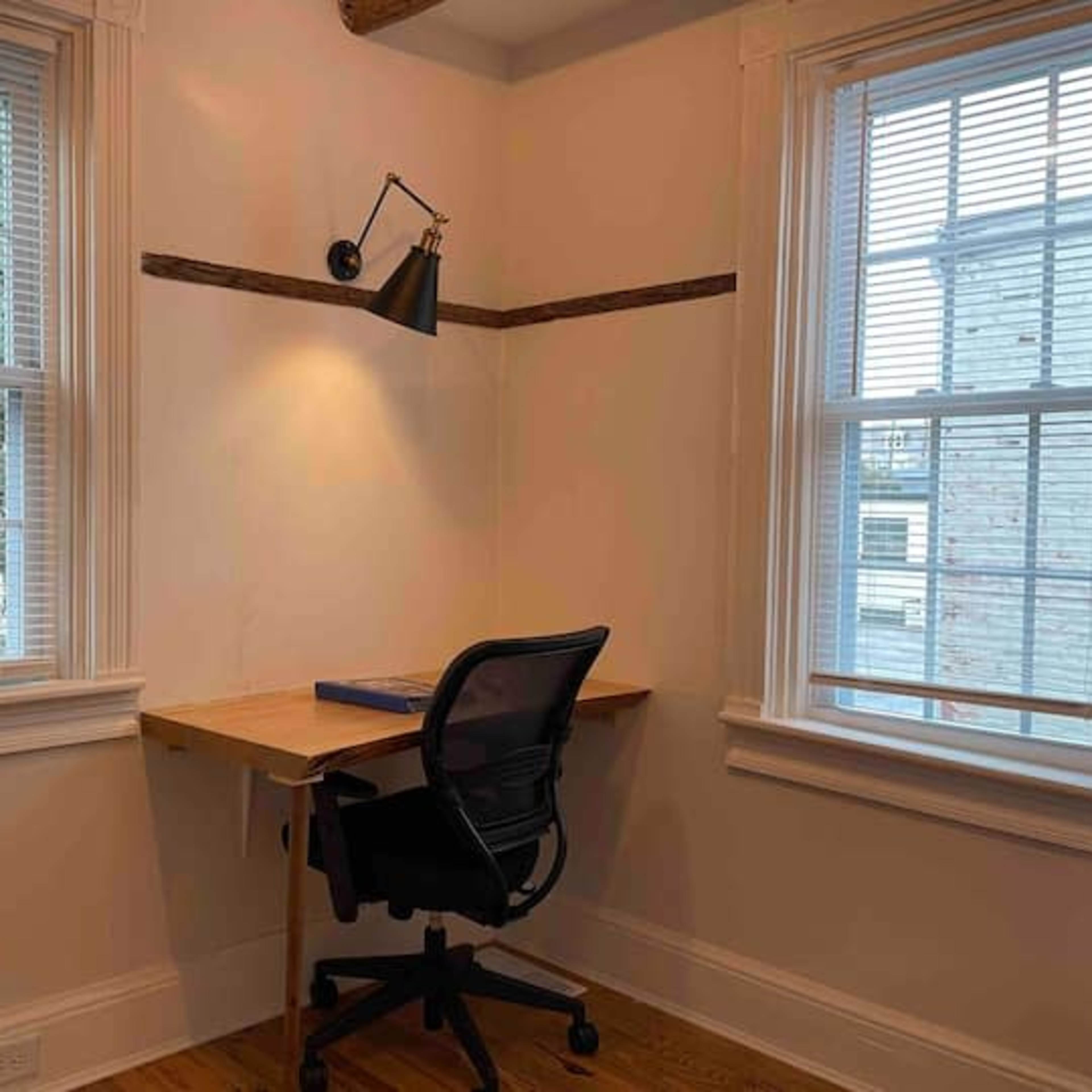 A desk and chair are positioned in a well-lit corner between two windows with wooden accents on the walls.