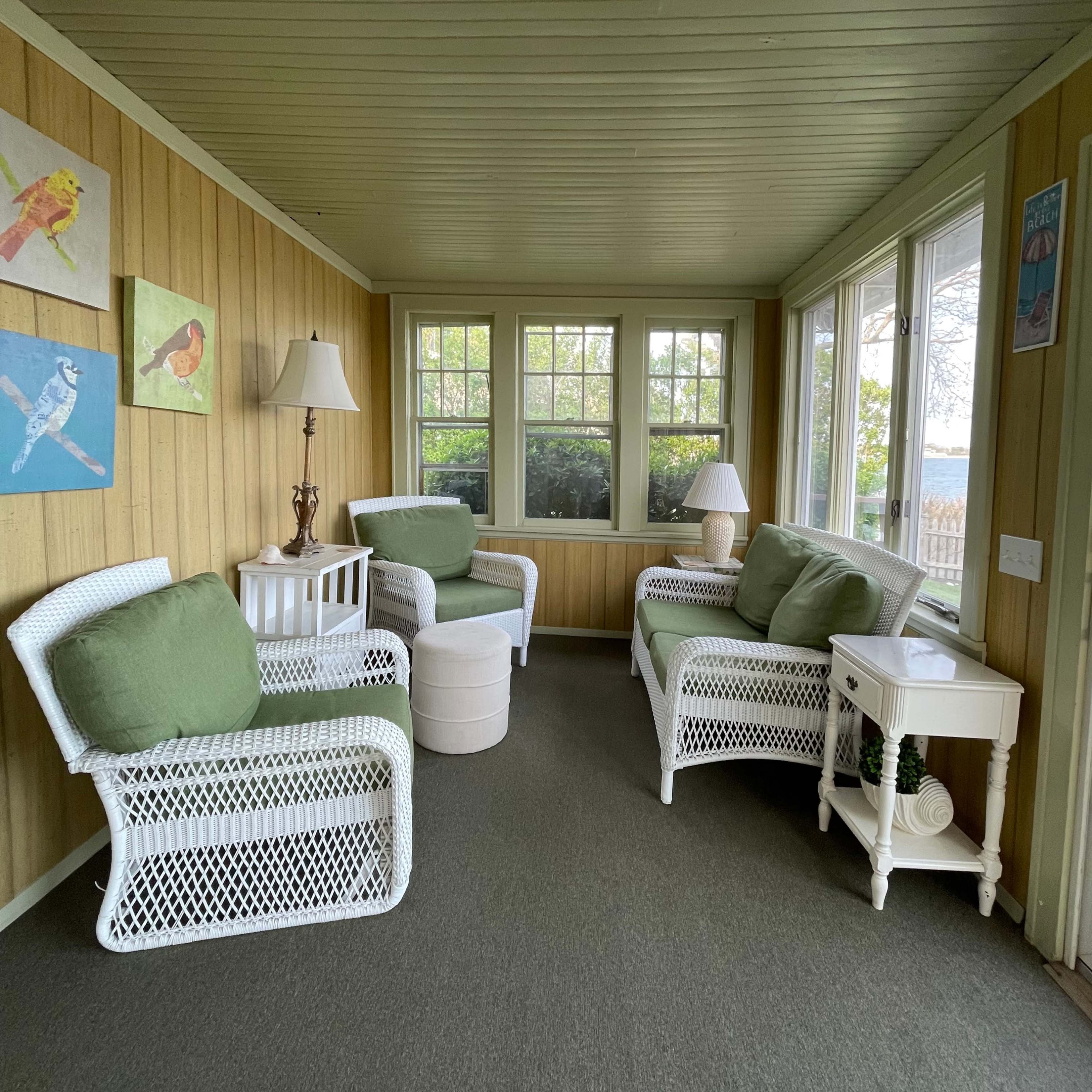 A sunroom with white wicker furniture, green cushions, and wooden walls, featuring windows that overlook an outdoor view.