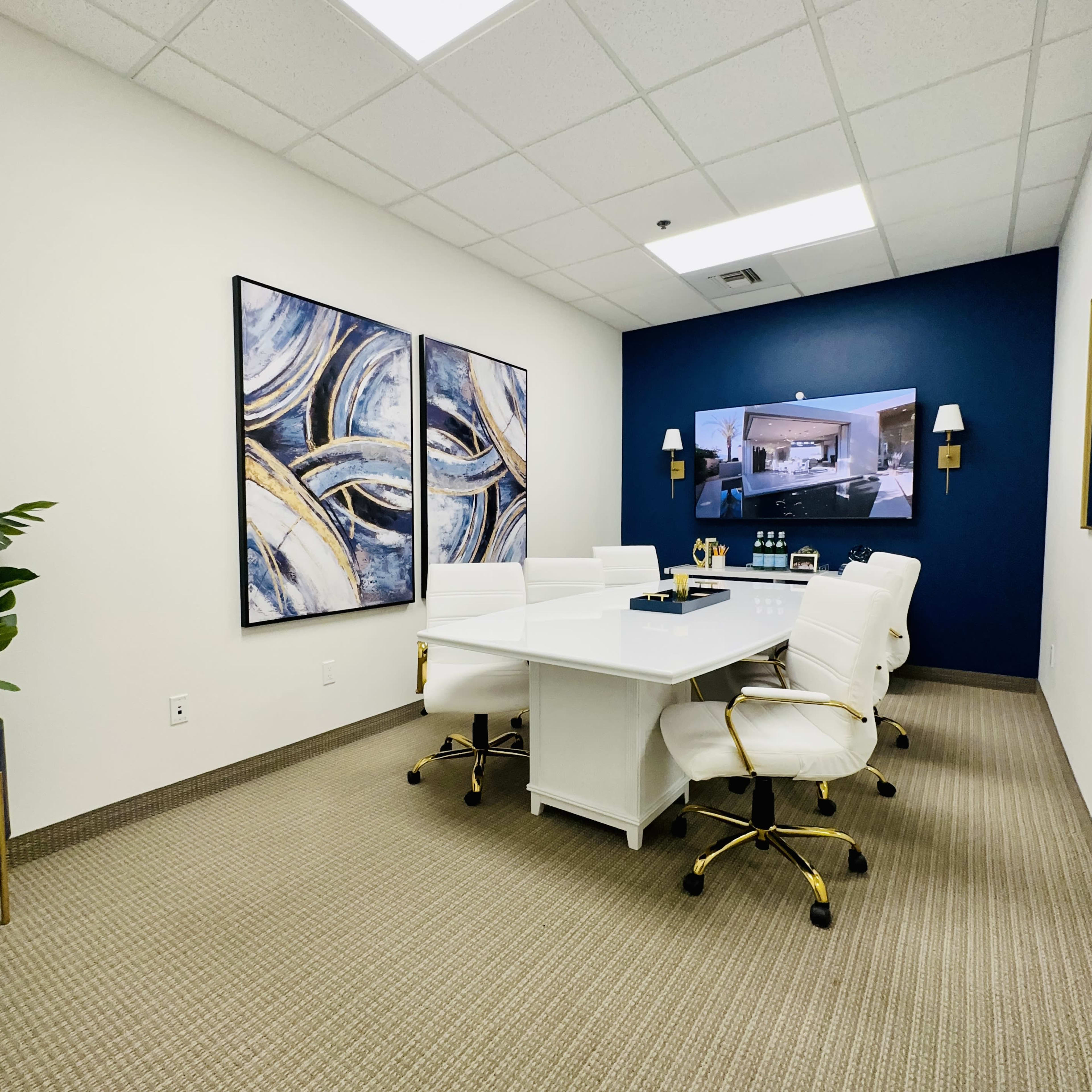 A modern conference room with a white table surrounded by white chairs, complemented by blue wall decor and a flat-screen monitor.