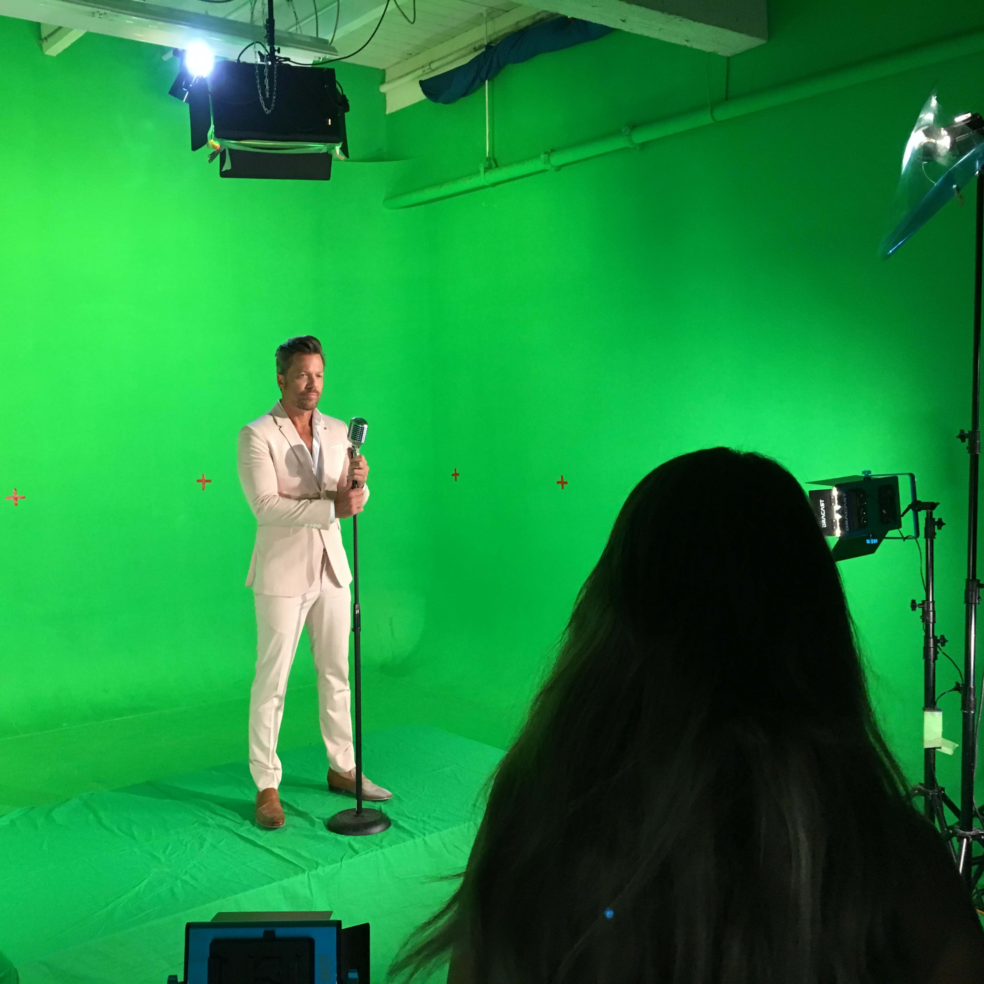 A man in a light-colored suit stands on a green background, holding a microphone, while a woman with long hair observes from behind a camera setup.