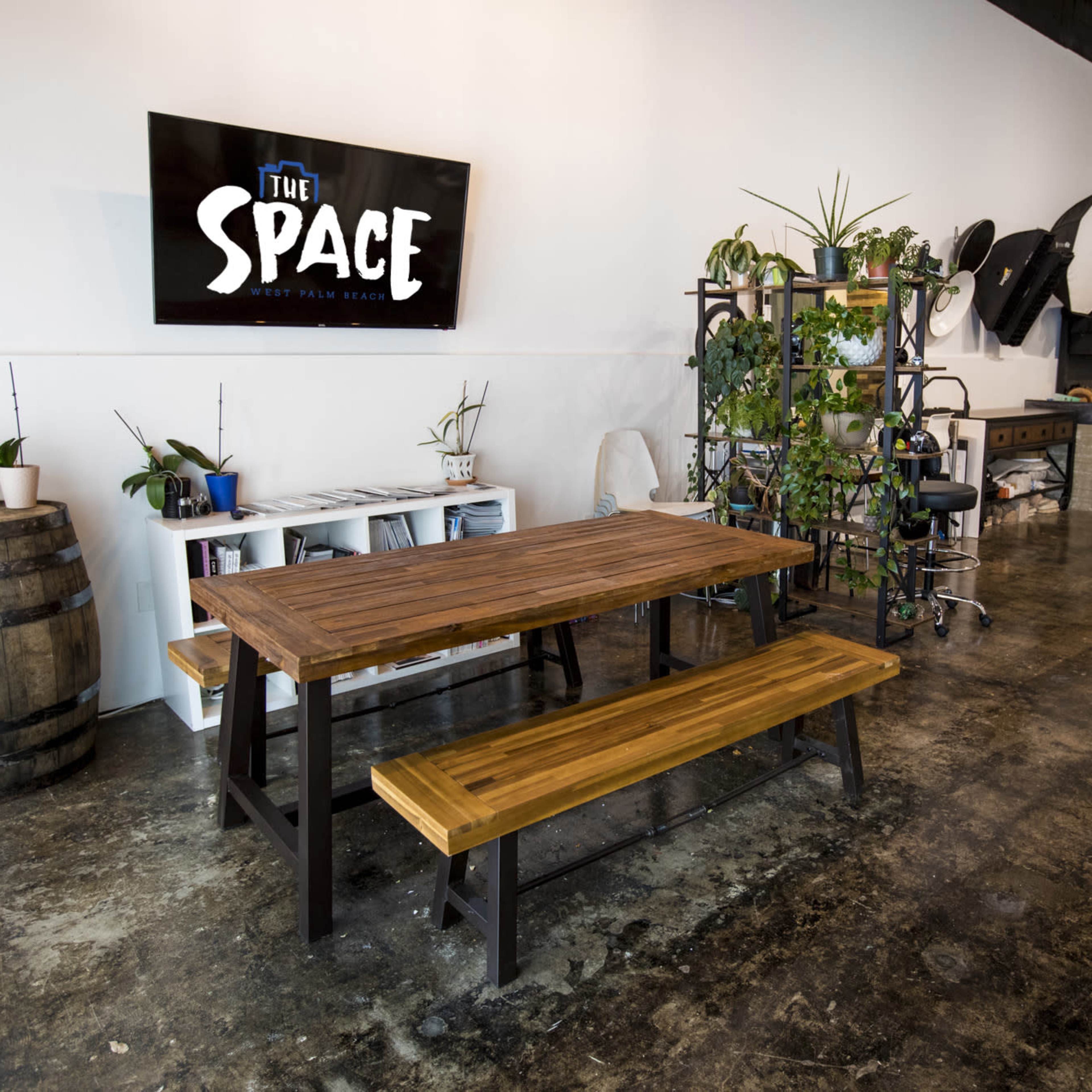 A spacious interior featuring a wooden dining table with benches, greenery on shelves, and a wall-mounted screen displaying the text "THE SPACE."