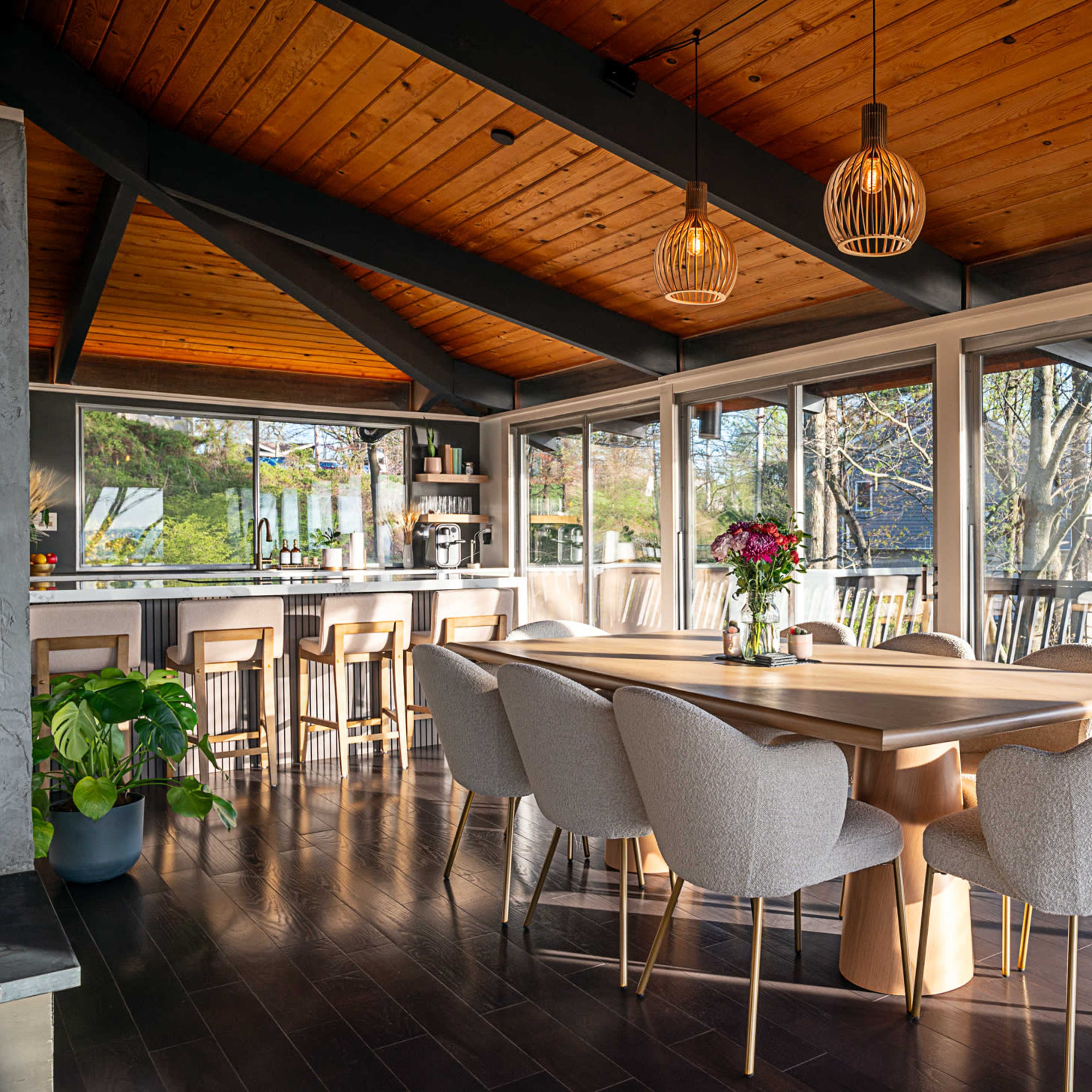 A modern dining area features a wooden table surrounded by upholstered chairs, with large glass windows that overlook a natural setting.