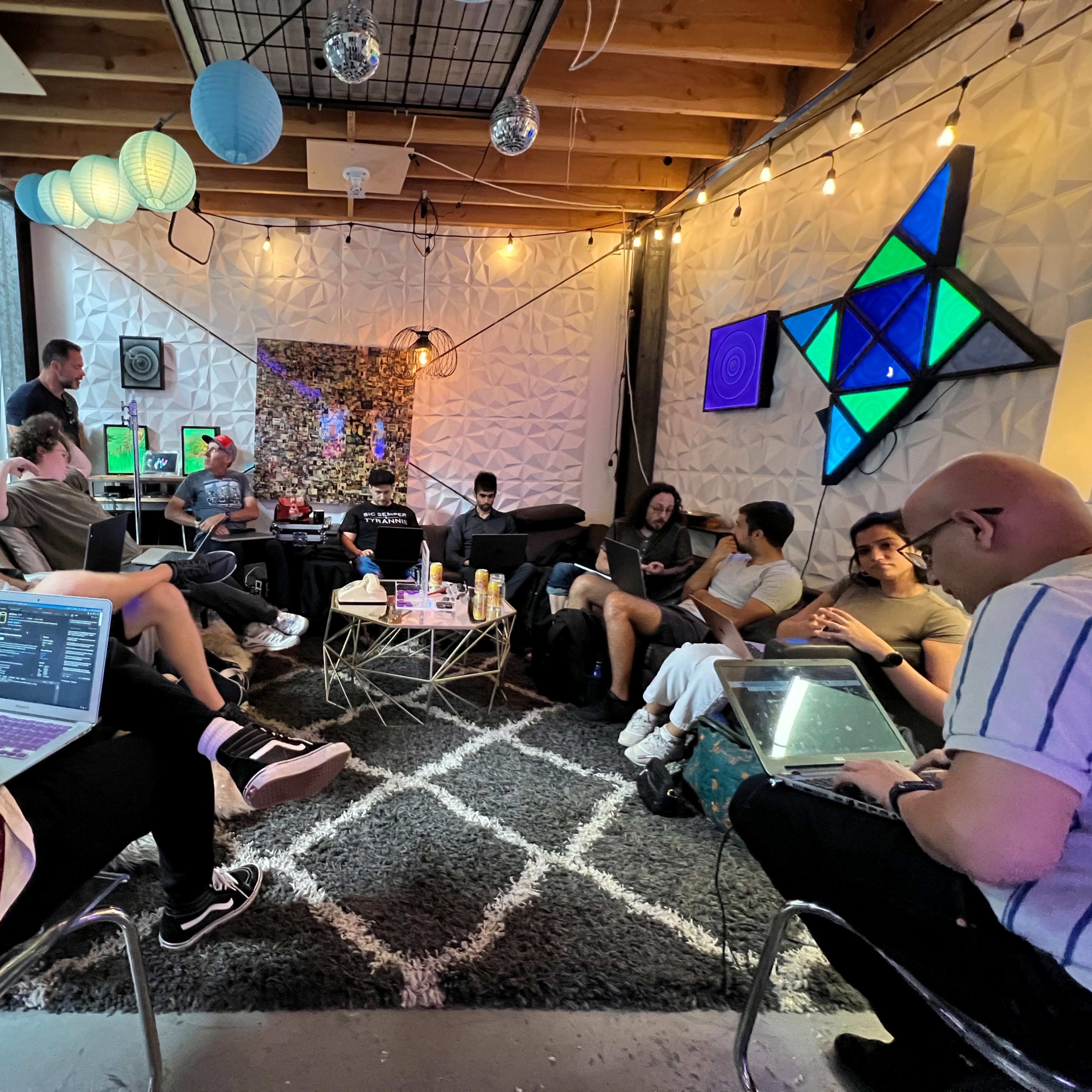 A group of people sit in a modern, well-decorated lounge, engaging in discussions and working on laptops while surrounded by colorful lighting and plants.