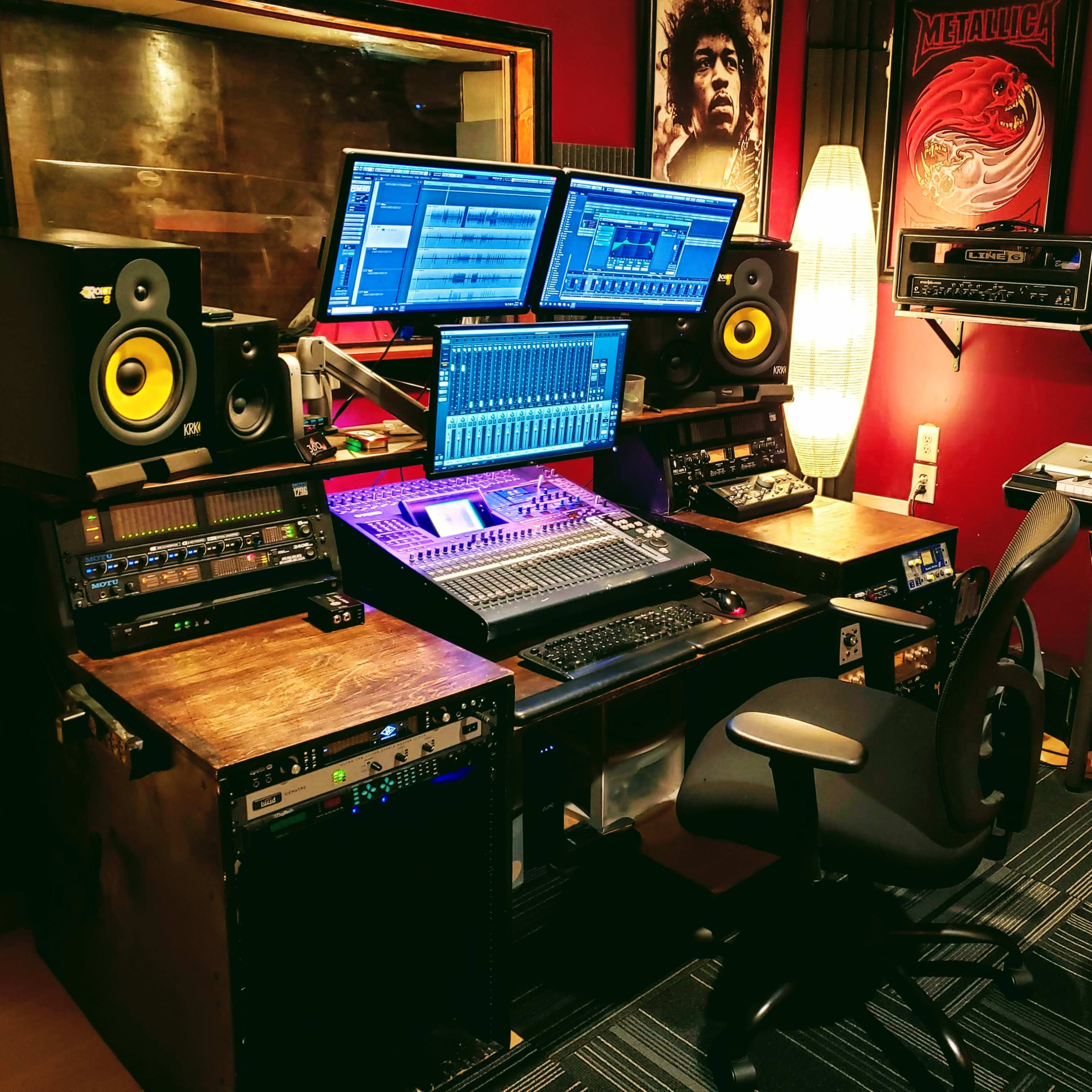 A music production studio with multiple screens, audio equipment, and a swivel chair arranged around a central mixing console.