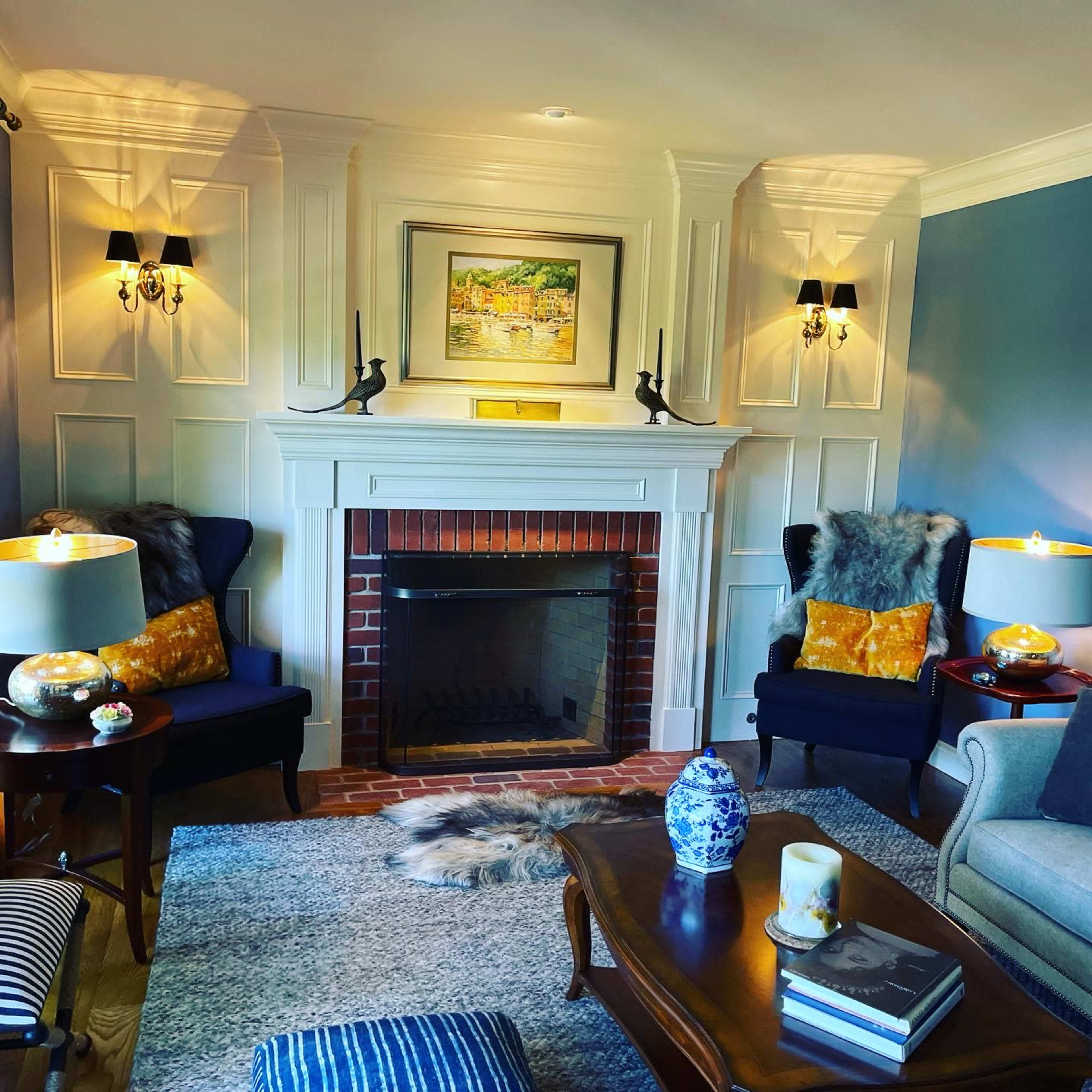 A cozy living room features a brick fireplace with a painting above it, surrounded by two armchairs and a coffee table, all illuminated by wall sconces.