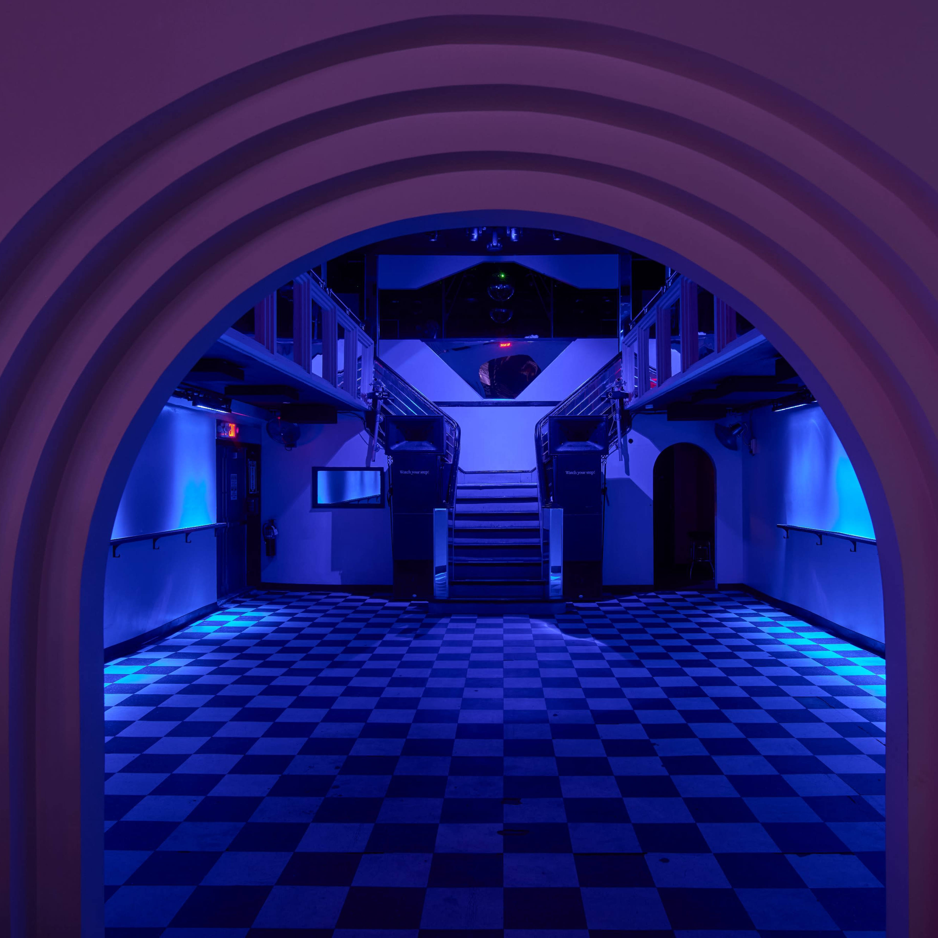 A dimly lit interior of a building with a checkered floor and an arched entrance leading to a staircase in the background.