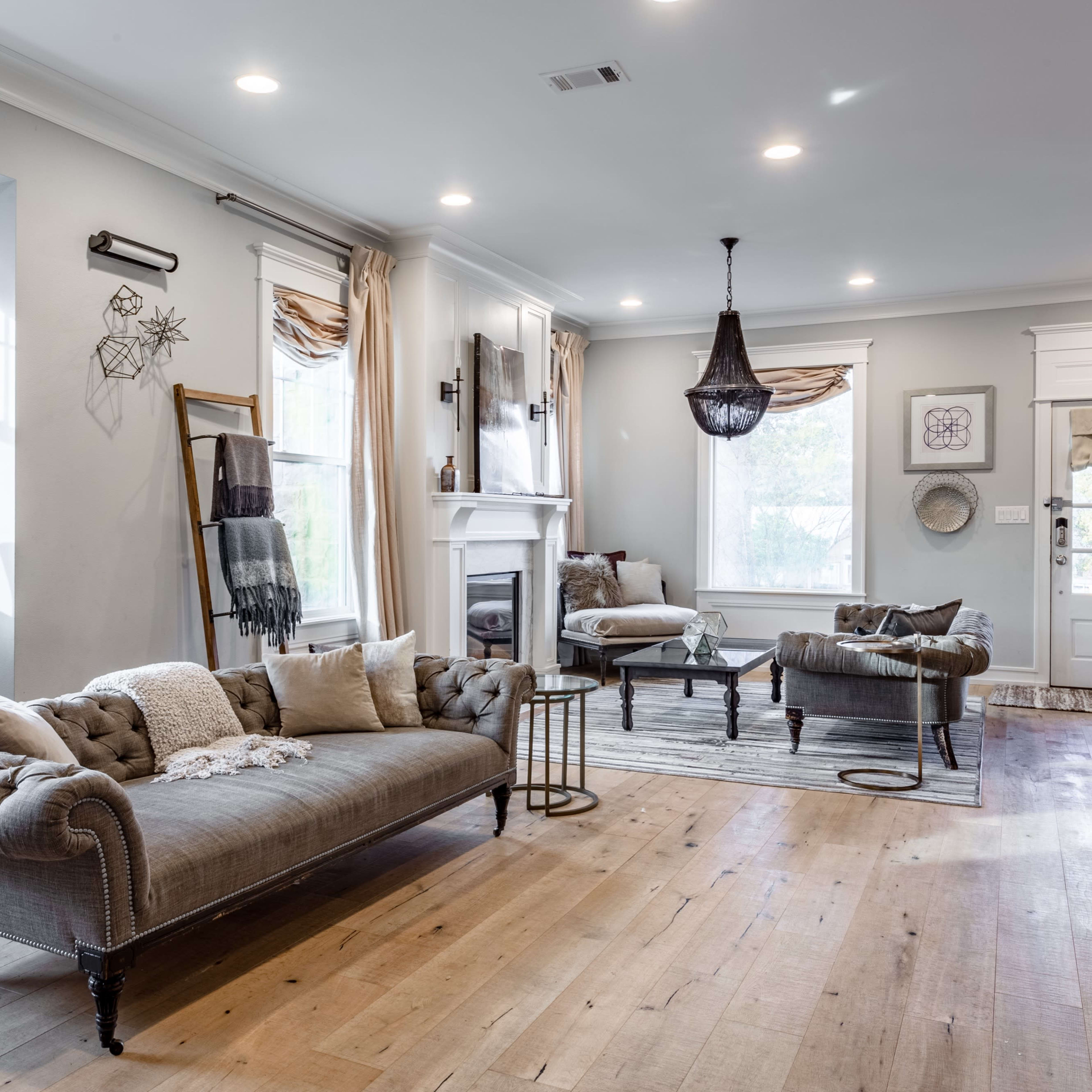 A living room features a tufted sofa, a chair, and a coffee table, all arranged around a fireplace with large windows allowing natural light.
