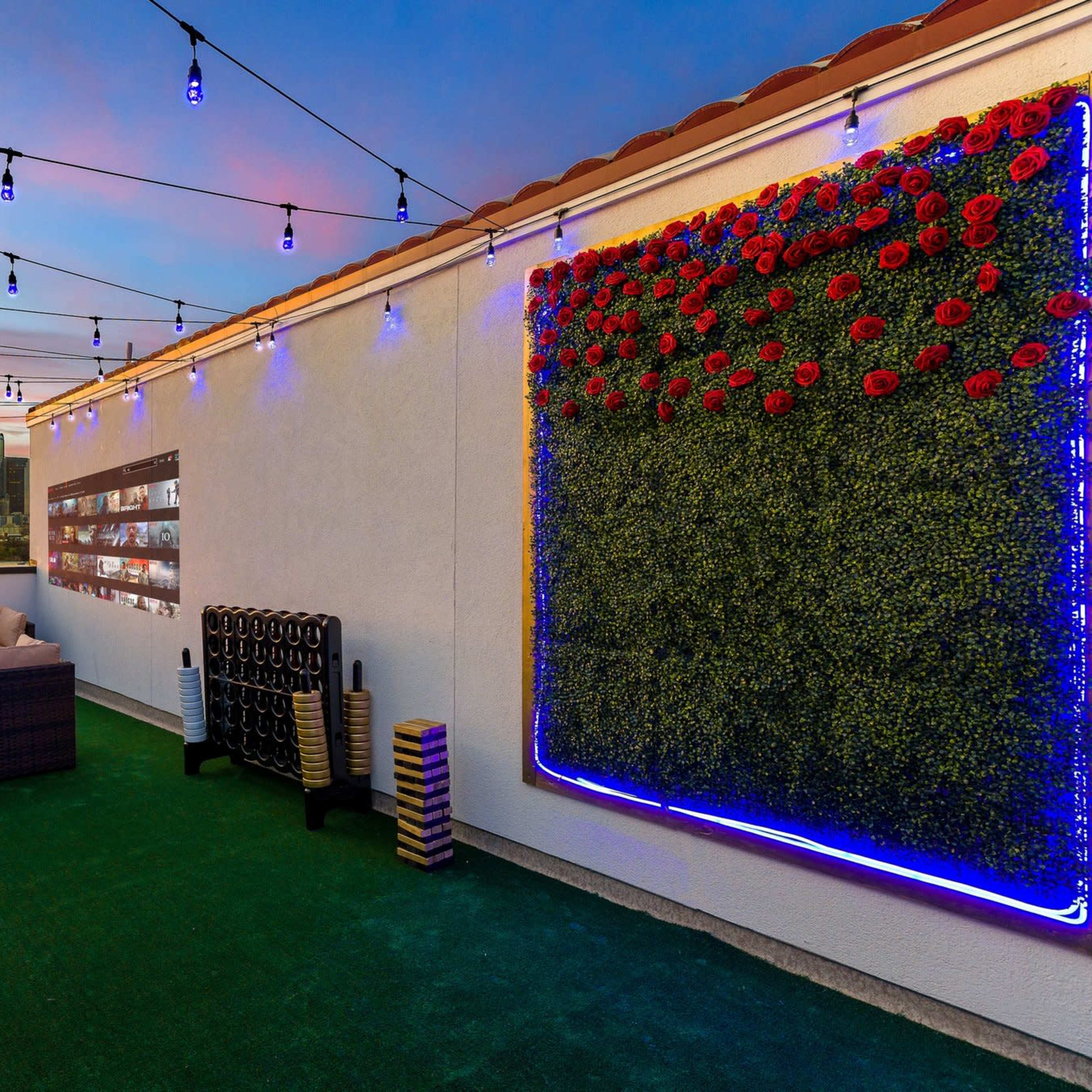 A rooftop lounge area features a couch, a vertical garden adorned with roses, and city skyline views under string lights at sunset.