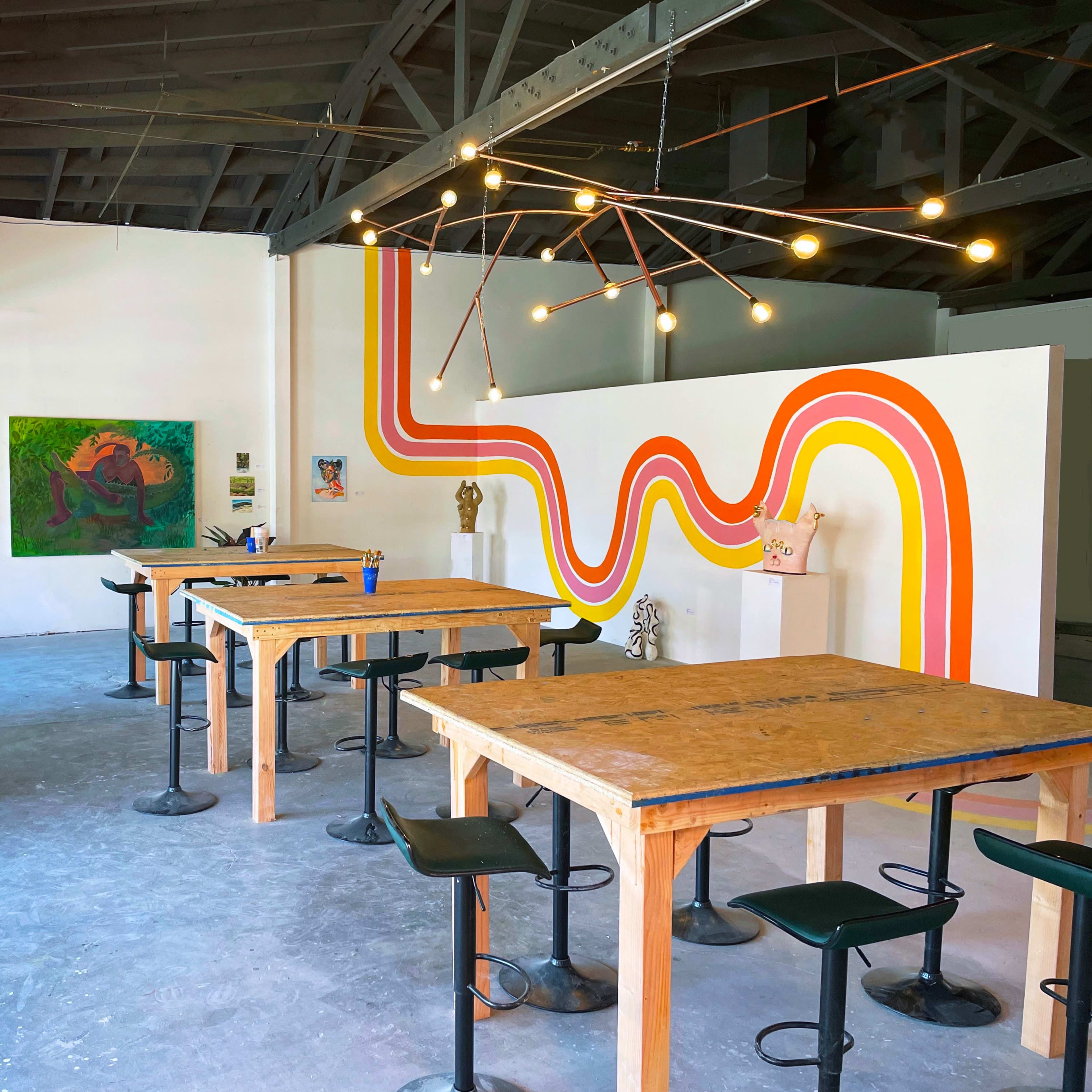 A spacious art studio with wooden tables and colorful wall art featuring bold orange and yellow lines.