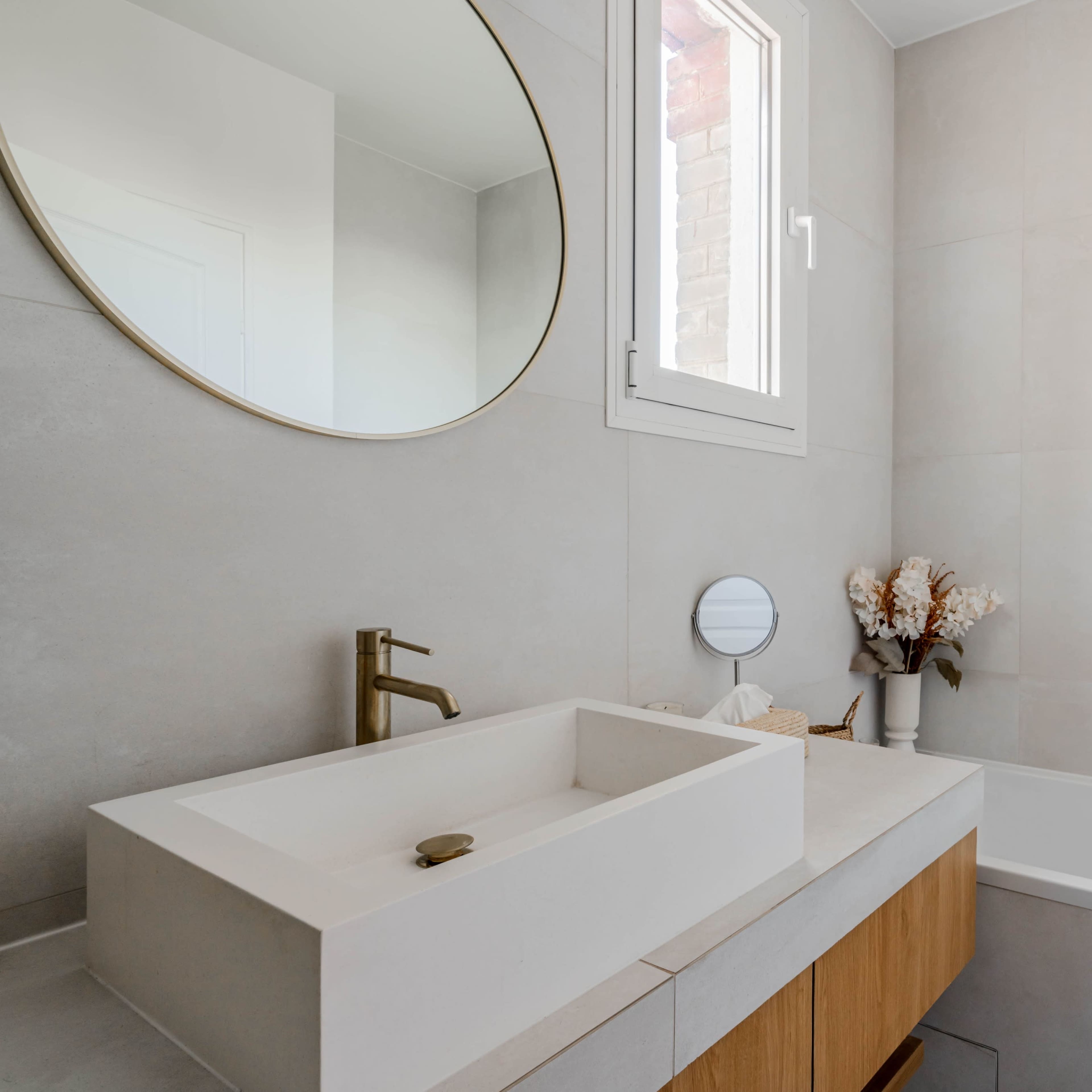 A modern bathroom features a rectangular sink with a gold faucet, a bathtub, and a round mirror, complemented by a decorative vase on a wooden cabinet.