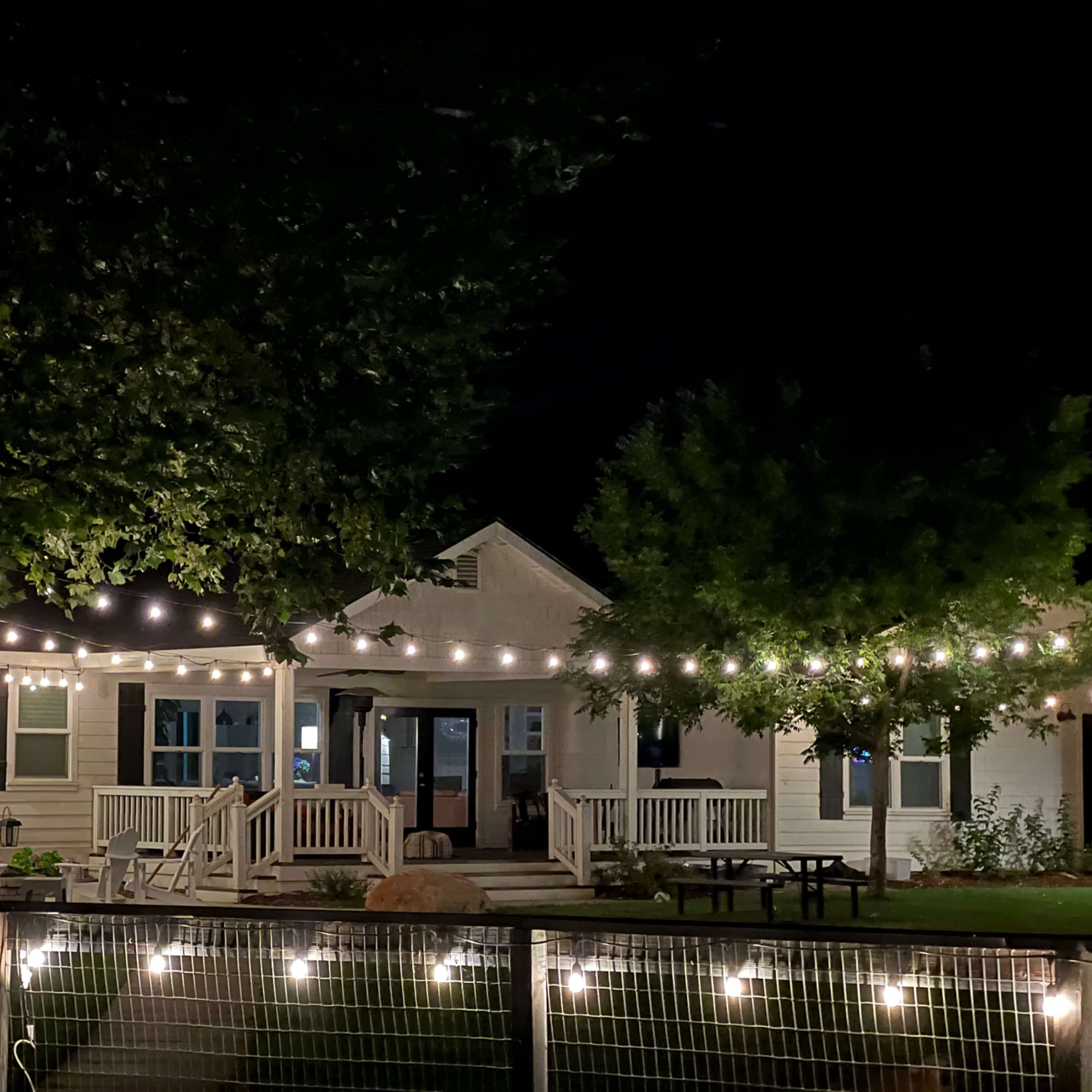 A well-lit house with string lights hangs over a patio area, surrounded by trees at night.
