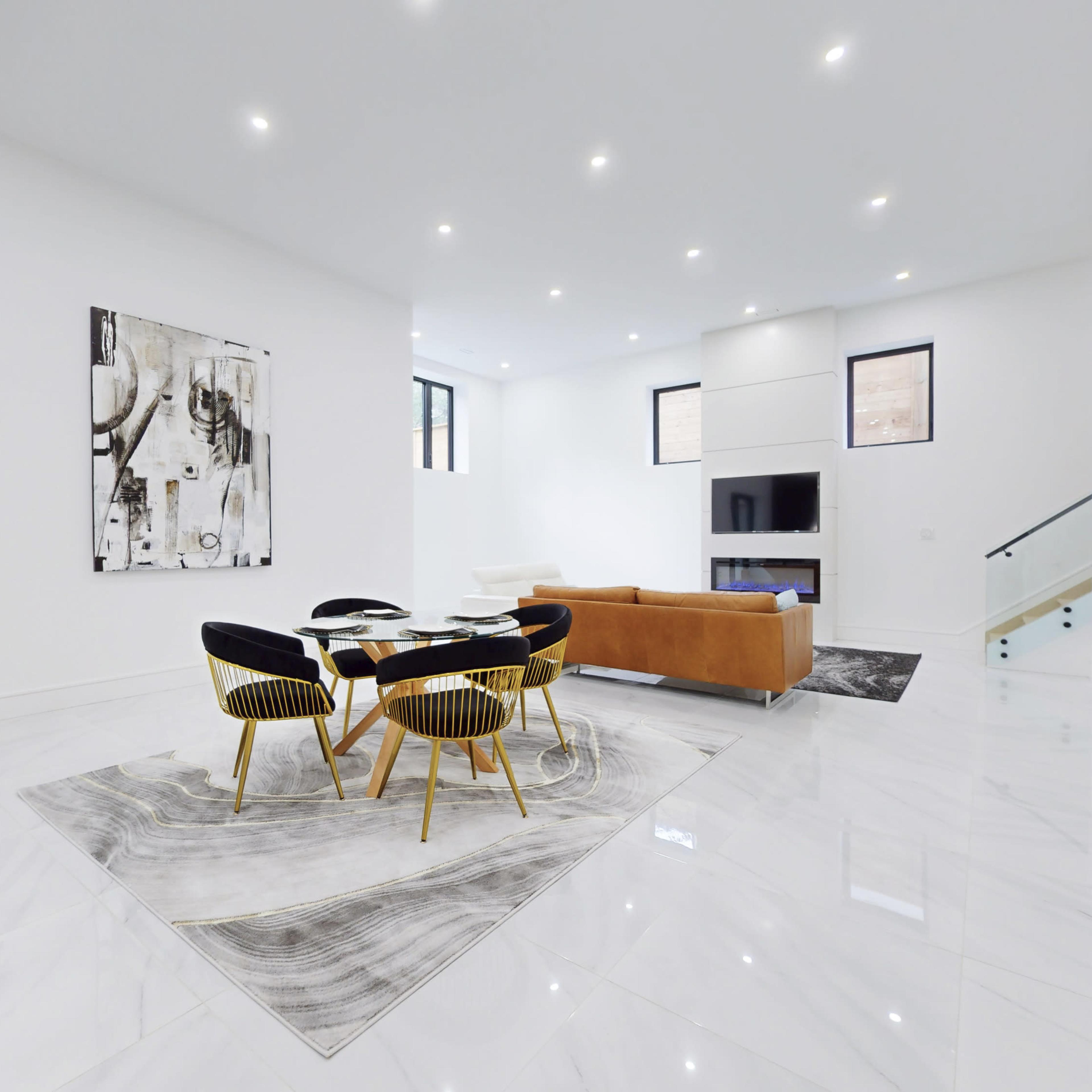 A spacious, modern living area features a round dining table surrounded by black and gold chairs, a large artwork on the wall, and a tan sofa.