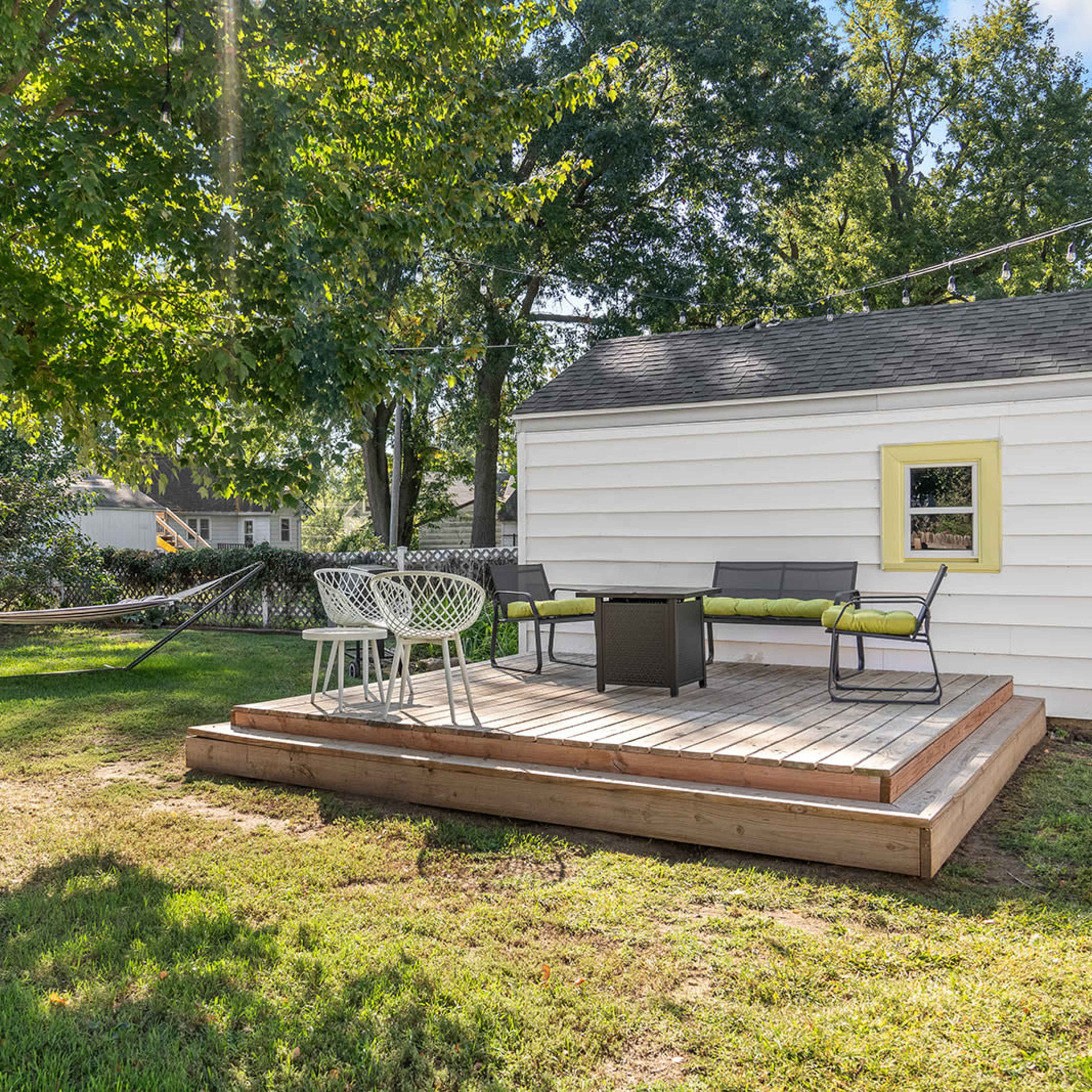 A wooden deck with seating and a table is situated beside a tree in a backyard, with a hammock visible in the background.